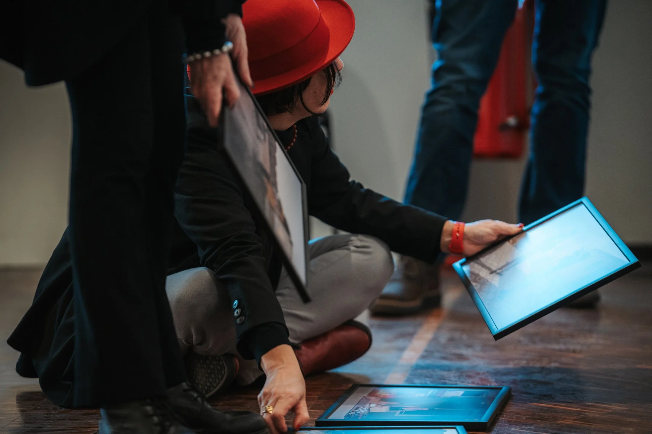 A woman wearing a red hard hat is crouching on the floor, holding a tablet. She is surrounded by other people, one of whom is standing and holding a picture in their hand. The scene appears to be indoors, possibly at an event or presentation.