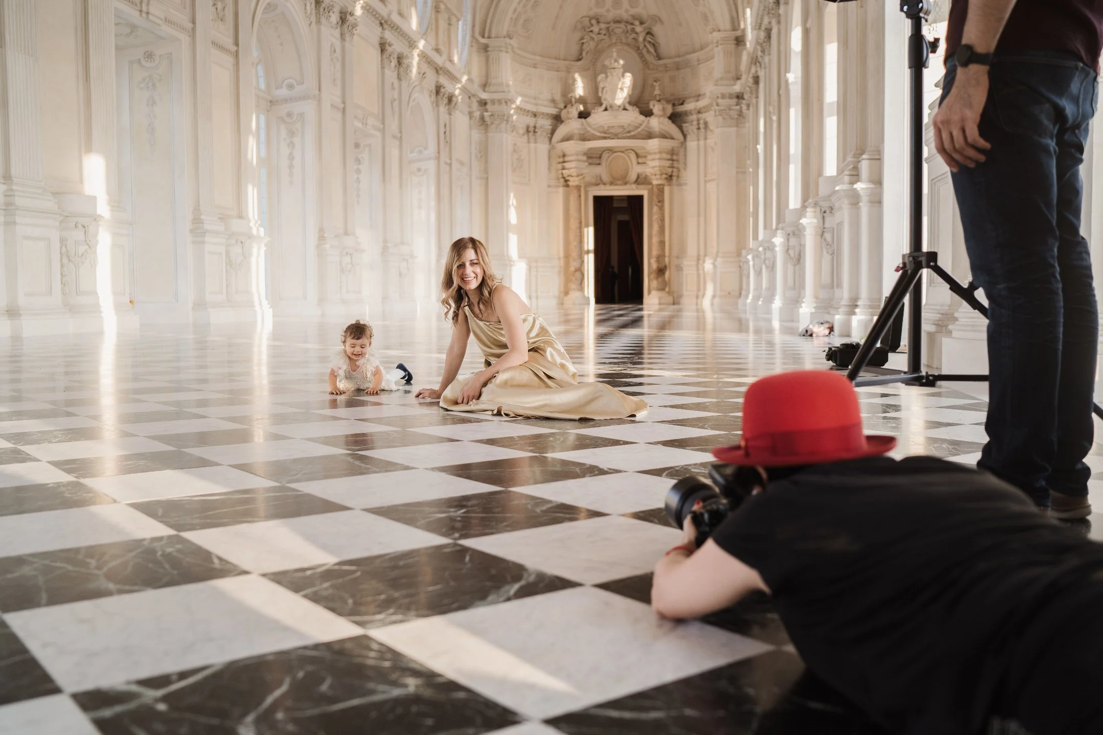 A woman and a young girl are sitting on a checkered marble floor inside a grand, ornate hall. The woman is smiling at the camera, while the girl is crawling. A photographer wearing a red hat is lying on the floor, taking pictures with a professional camera. There are additional people and equipment in the background.