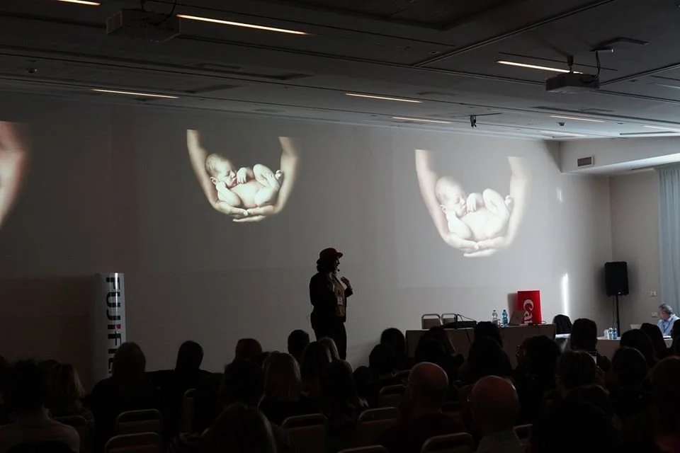 Presentation in a conference room with a speaker standing in front of a projection of a baby's hands holding an older child's feet, and an audience watching.