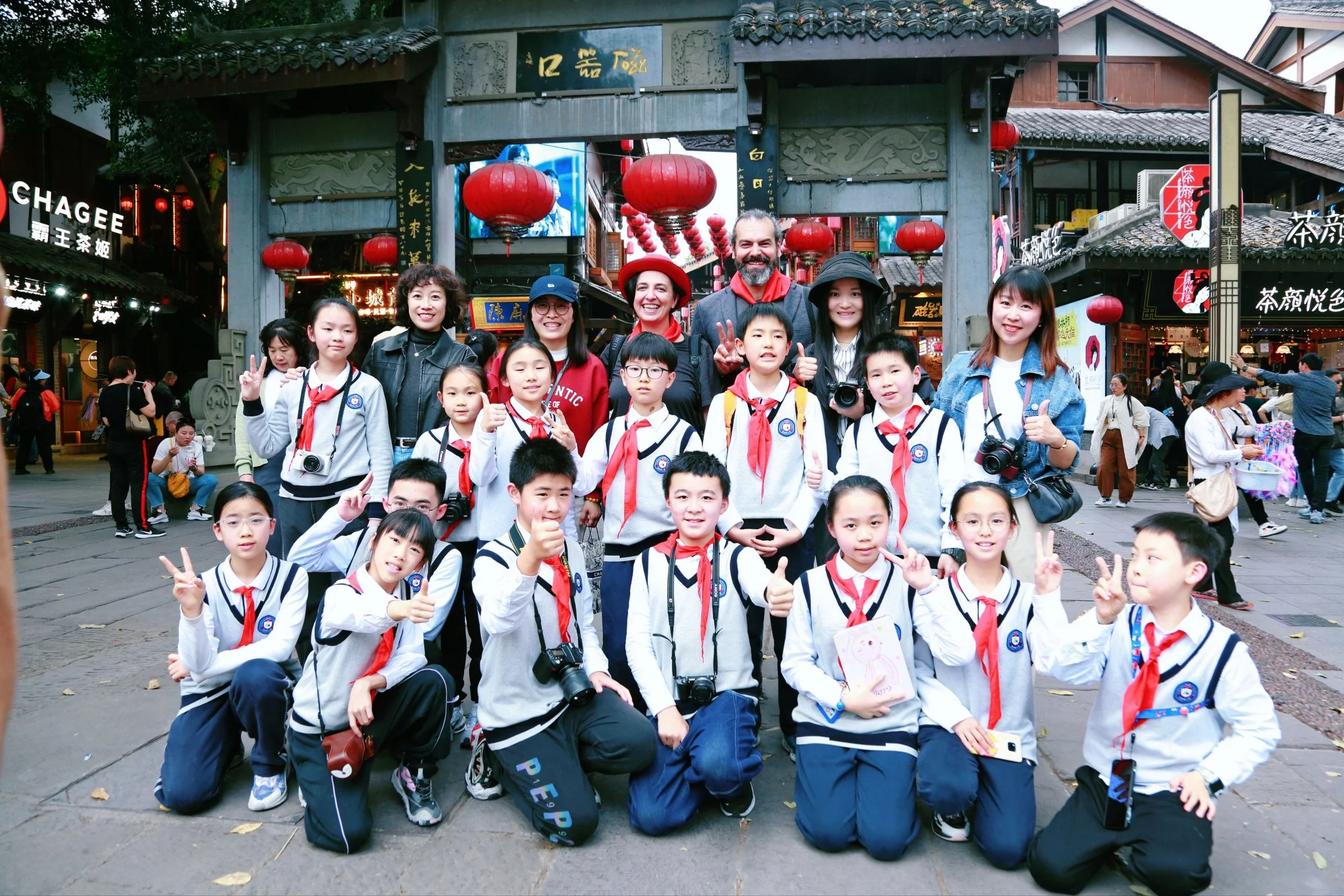 A large group of children and adults posing for a photo in front of a traditional Chinese entrance decorated with red lanterns. The children are wearing school uniforms and some are holding cameras, while the adults are smiling and making peace signs.