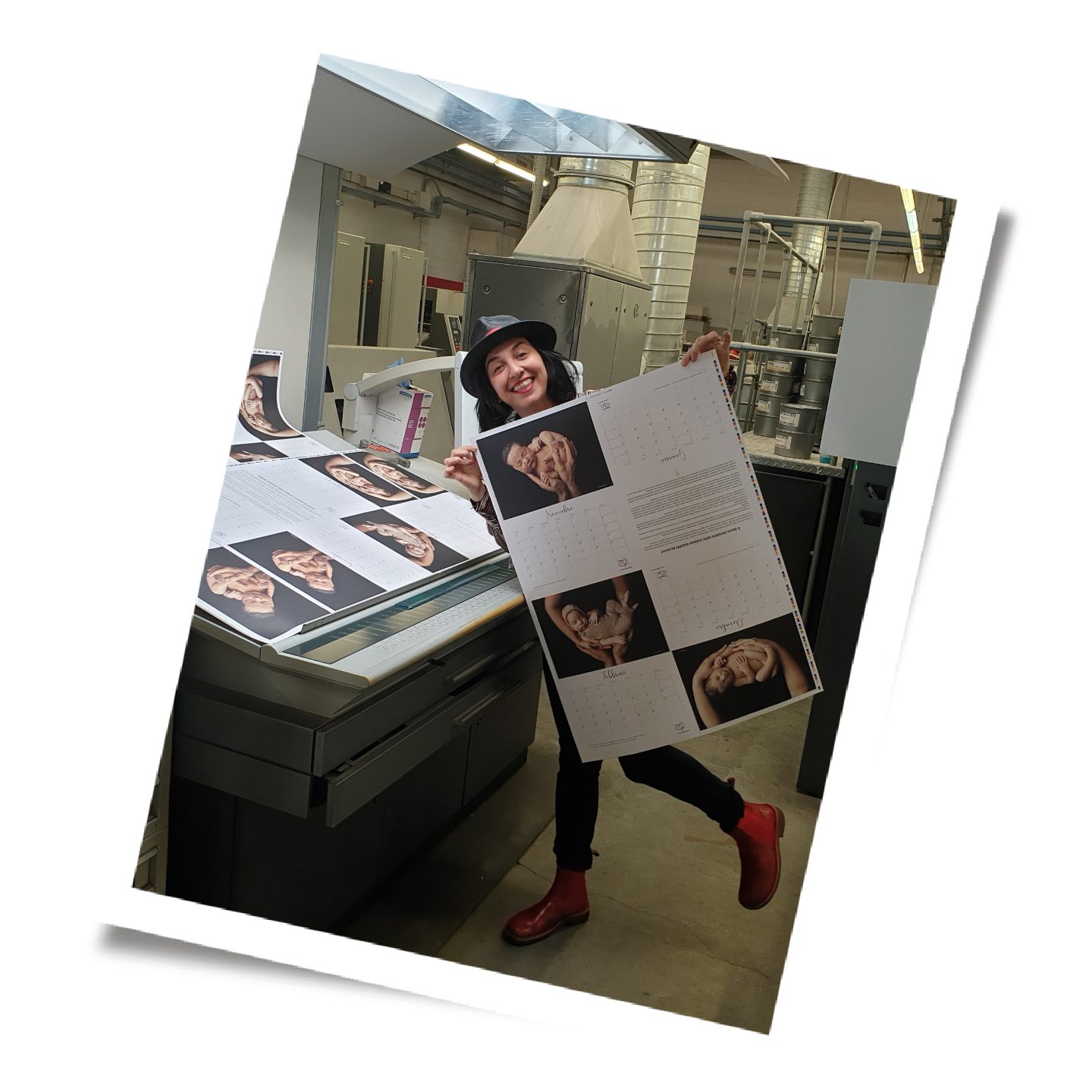 A woman holding a large calendar with photos of a man and woman in an industrial printing room, standing near printing equipment on a concrete floor.