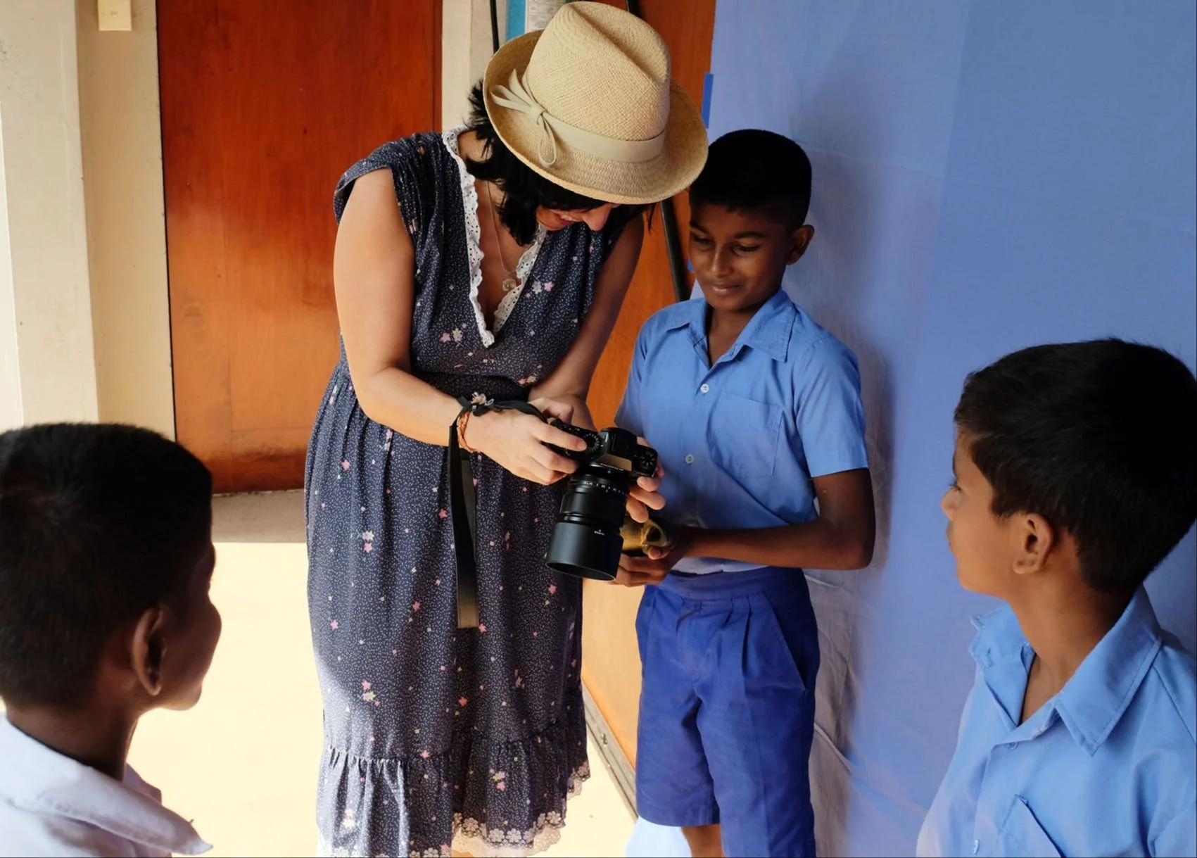 A woman wearing a summer dress and a wide-brimmed hat showing a camera to a young boy in a blue school uniform. Two other boys in similar uniforms are watching the scene, standing against a blue backdrop in a classroom or school setting.