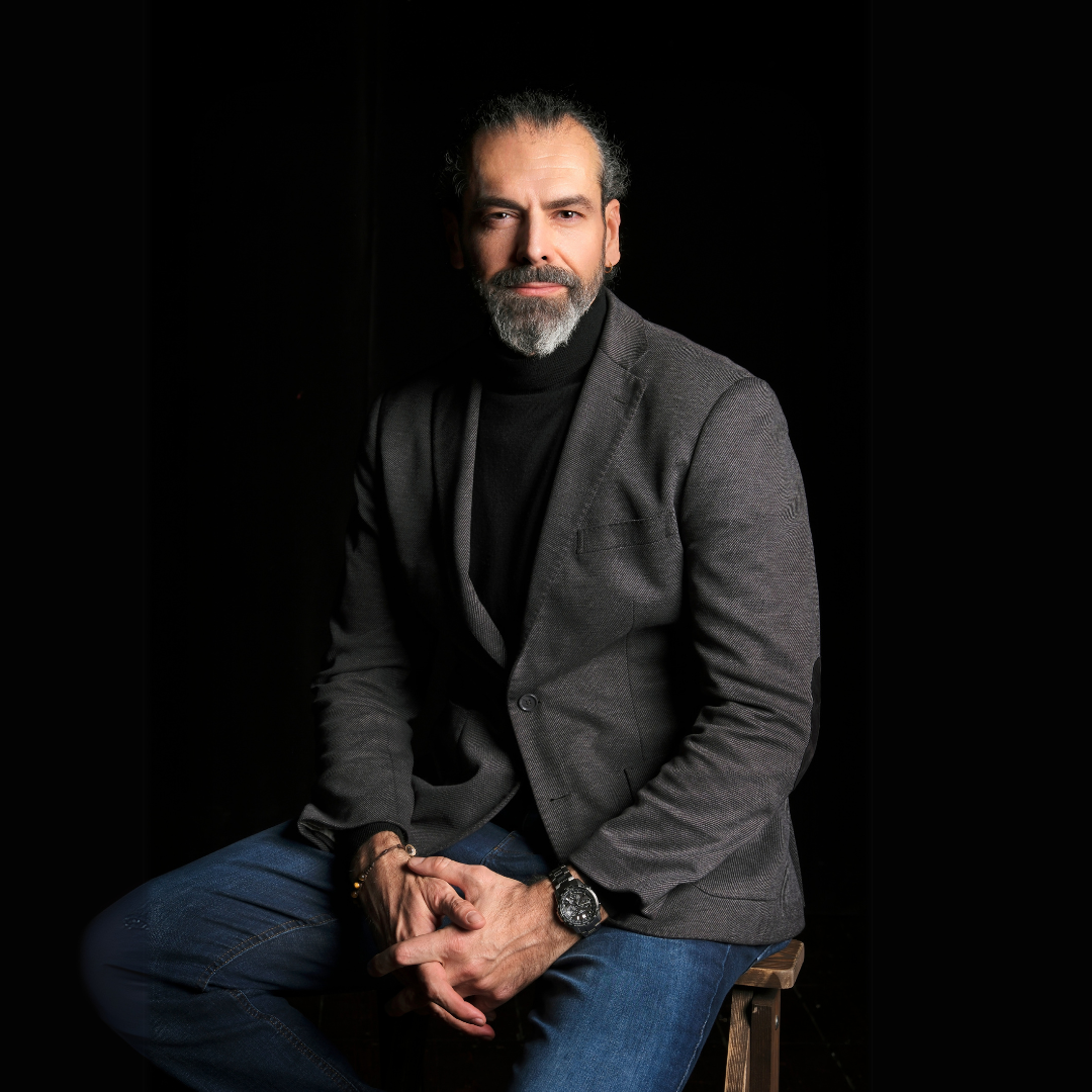 Andrea, Elena's husband, a man with a beard and dark hair, wearing a gray blazer and black turtleneck, sitting on a stool against a black background.