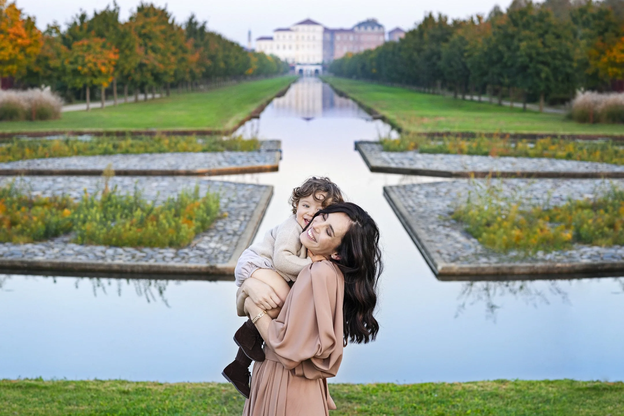 A woman holding a smiling child on her shoulder in a park with a water feature and a building in the background.