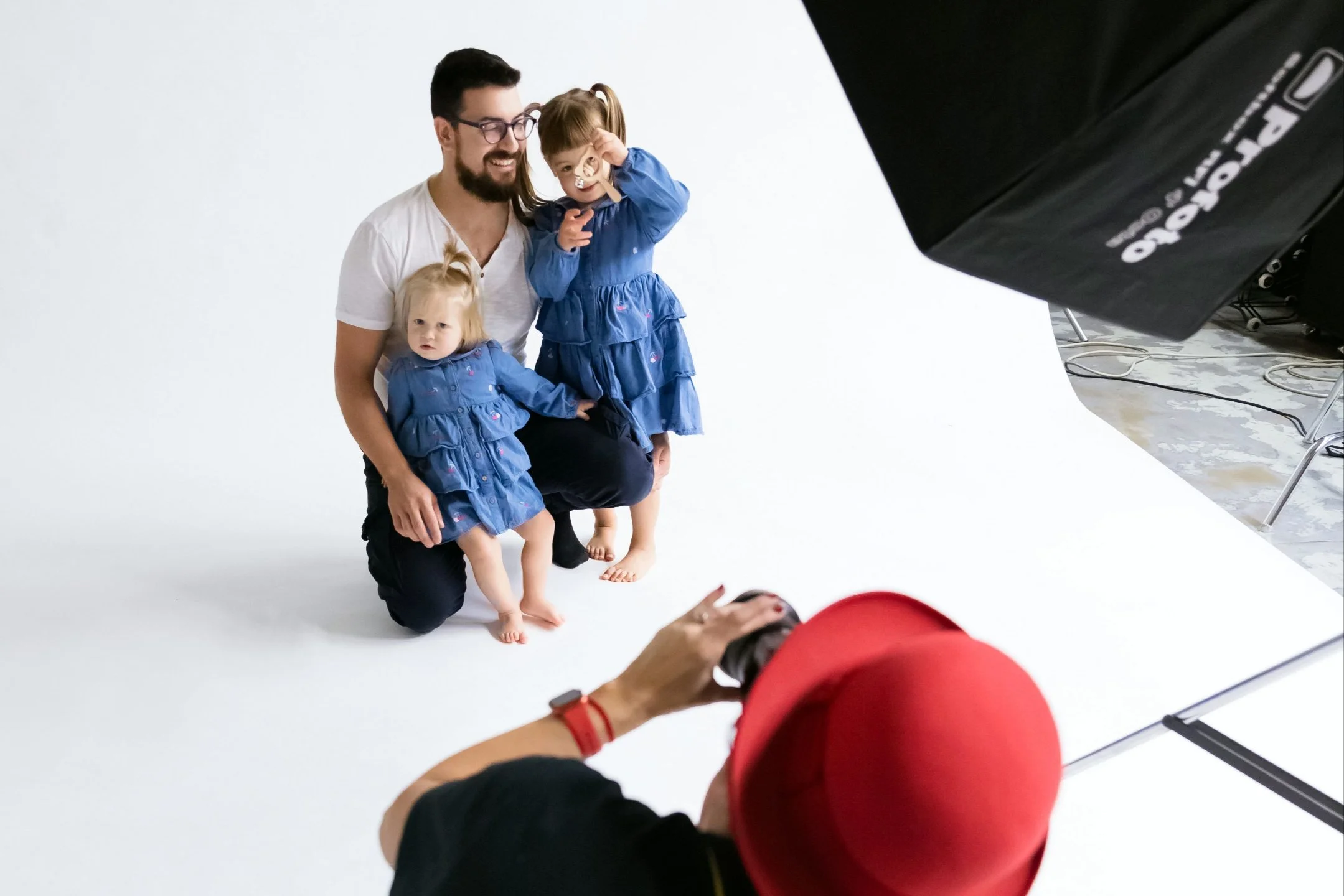 A man with two young girls, all dressed in matching blue dresses, pose for a photo in a studio with a white background. The man kneels while the girls stand, one of them making a gesture with her hand. The photographer wears a red hat and is taking the picture.