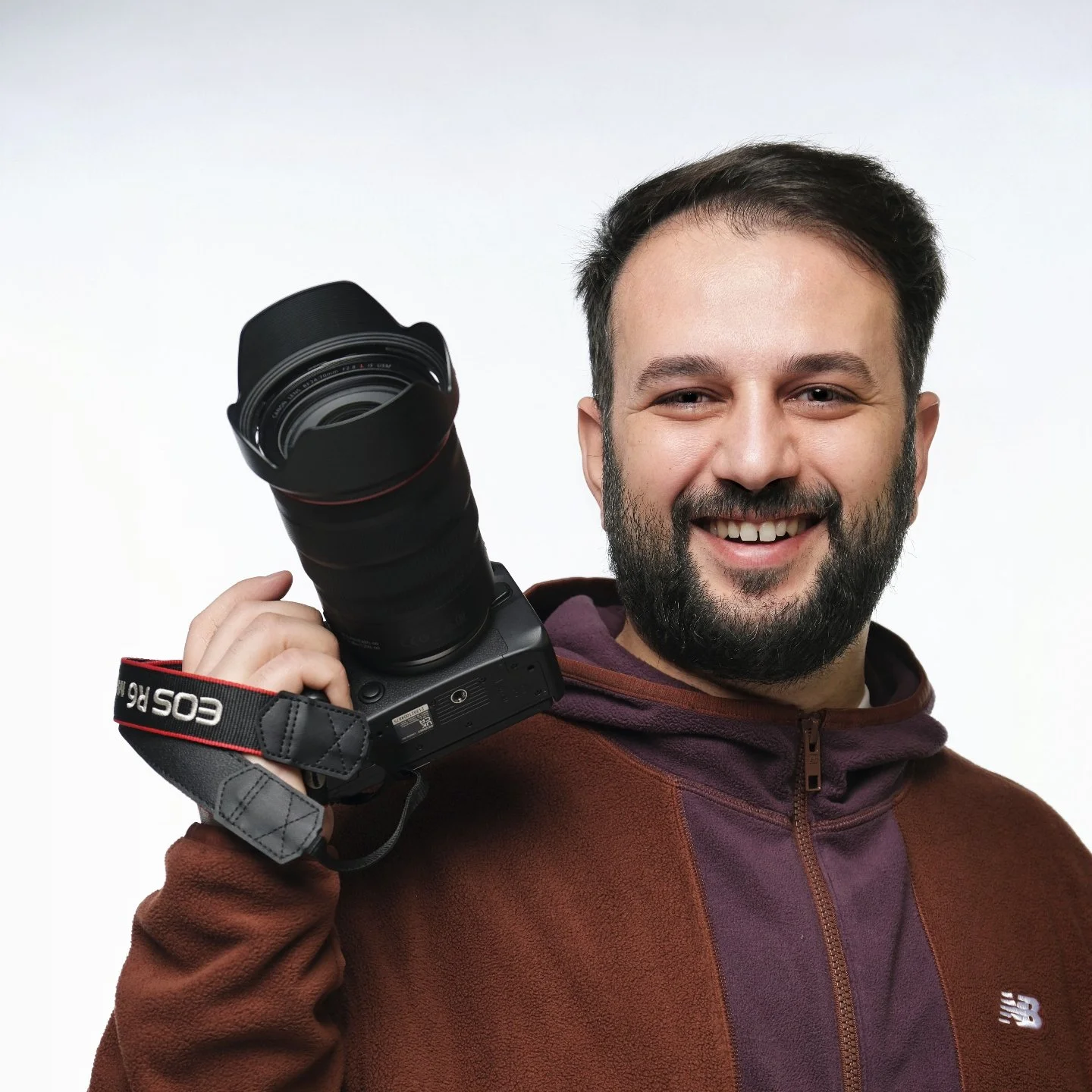 A man with a beard and short hair smiling while holding a camera with a large lens over his shoulder.