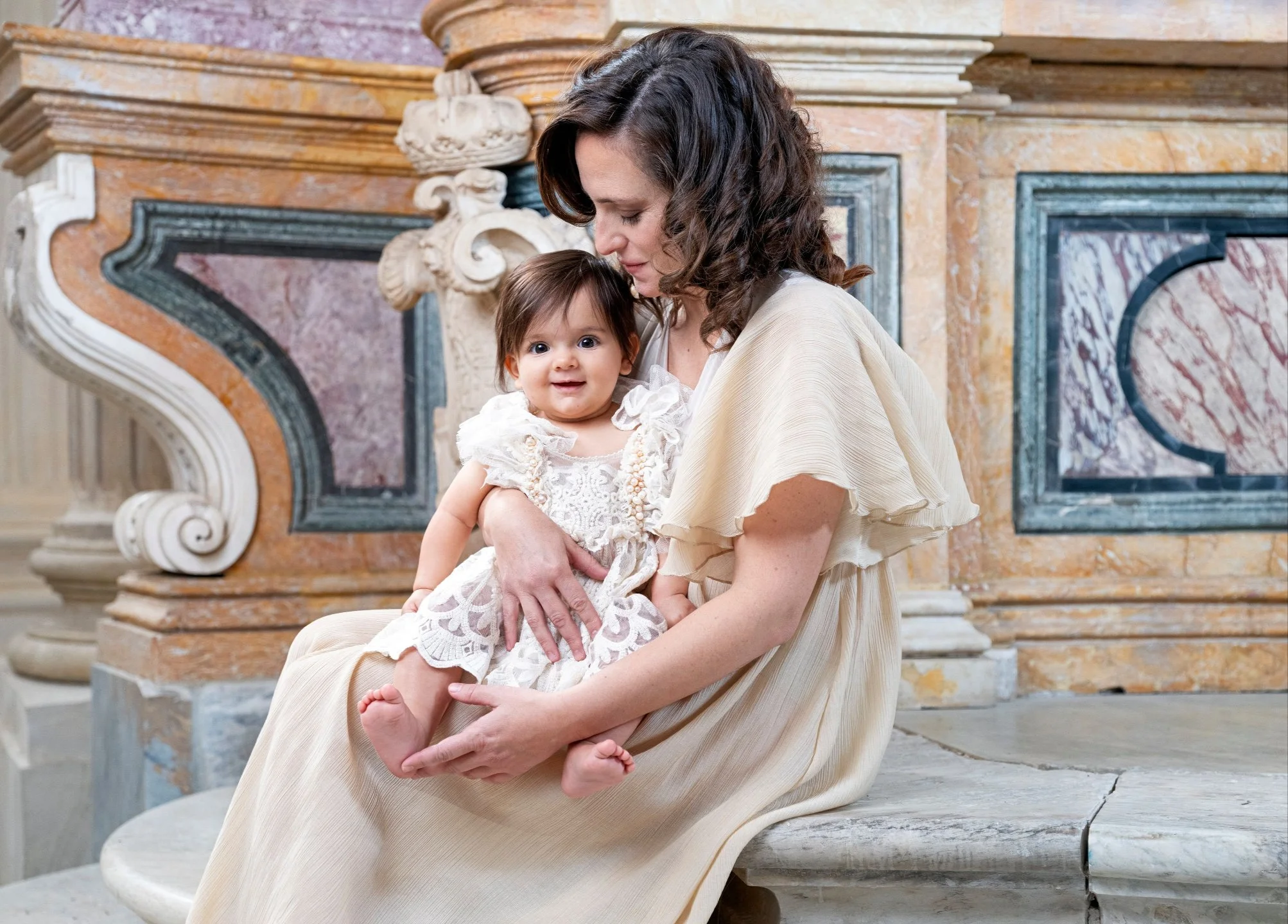 A woman with dark curly hair dressed in a cream dress holds a smiling baby girl with dark hair and big eyes, sitting on her lap on a stone bench in front of ornate marble decorations.