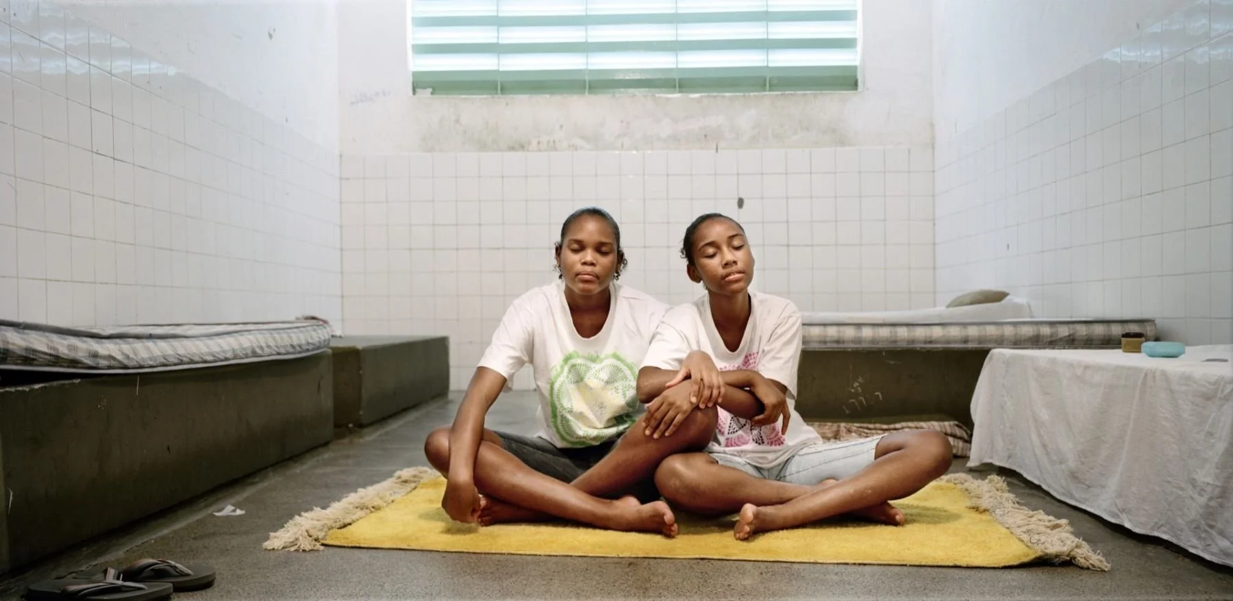 Two young girls sit cross-legged on a small yellow rug in a plain, white-tiled room, eyes closed, appearing to meditate or rest. There are twin beds with gray mattresses and some clothes or objects on a table to the side.