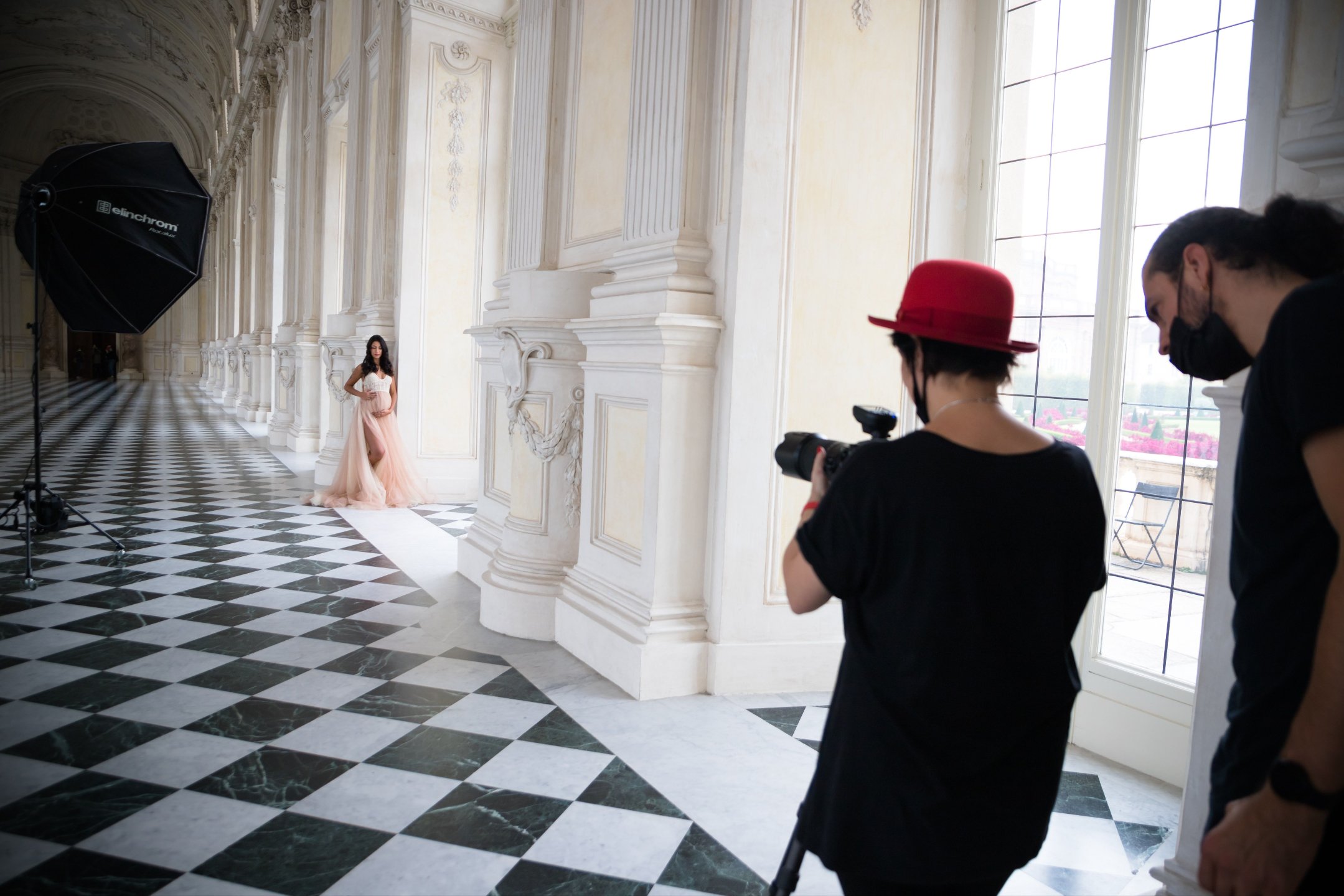Photoshoot in a grand, bright hall, a woman in a pink dress poses near tall windows with photographers capturing her image.