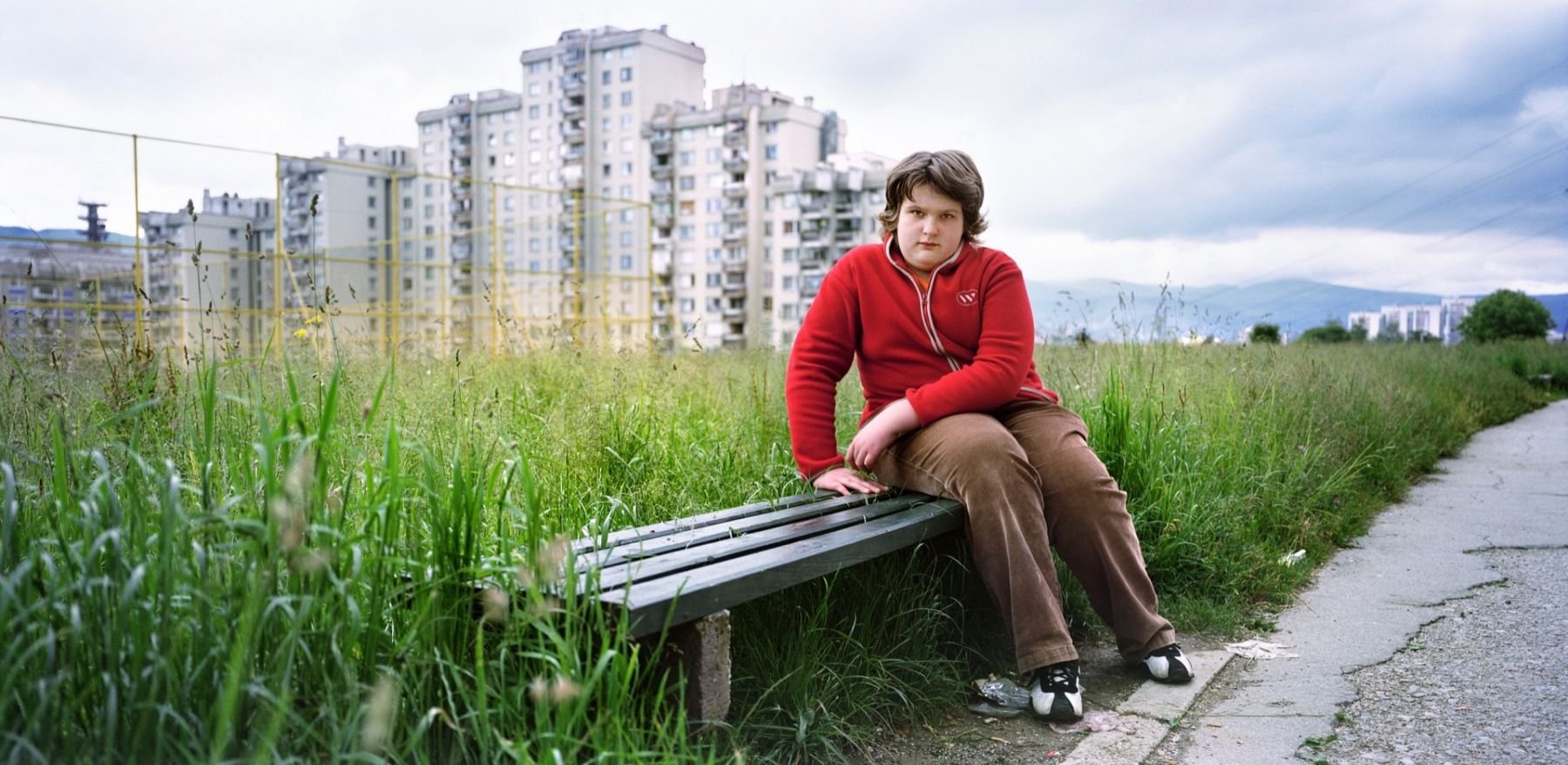 A young person sitting on a park bench next to a grassy area, with tall grass and yellow wildflowers, in an urban setting with high-rise buildings in the background on a cloudy day.