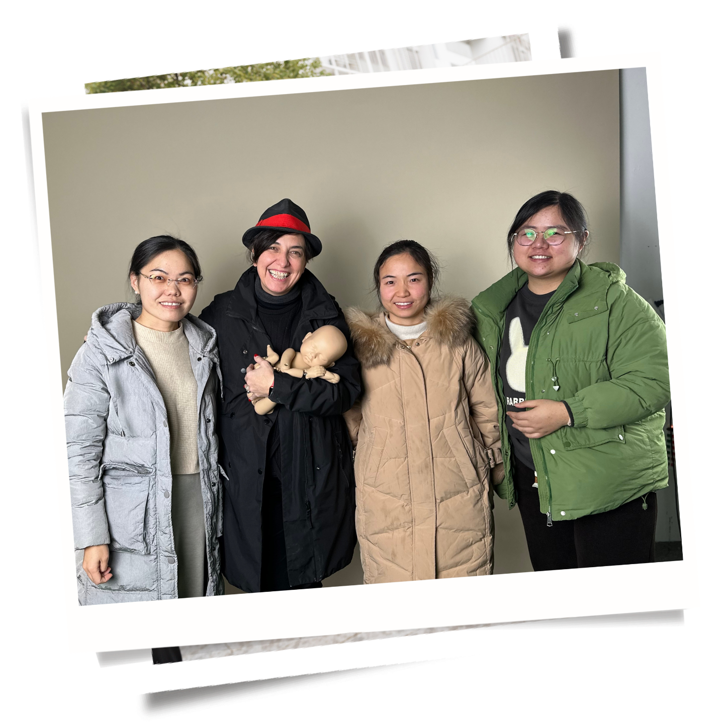 Four women standing together indoors, with one woman holding baby dolls, all smiling at the camera.