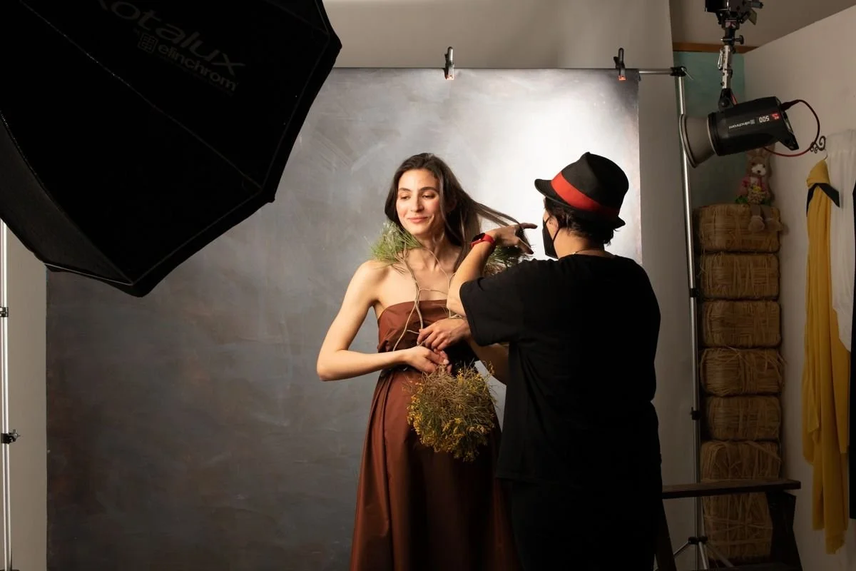 A woman in a brown dress is posing during a photoshoot with a photographer adjusting her hair or headpiece inside a studio with professional lighting and a gray backdrop.