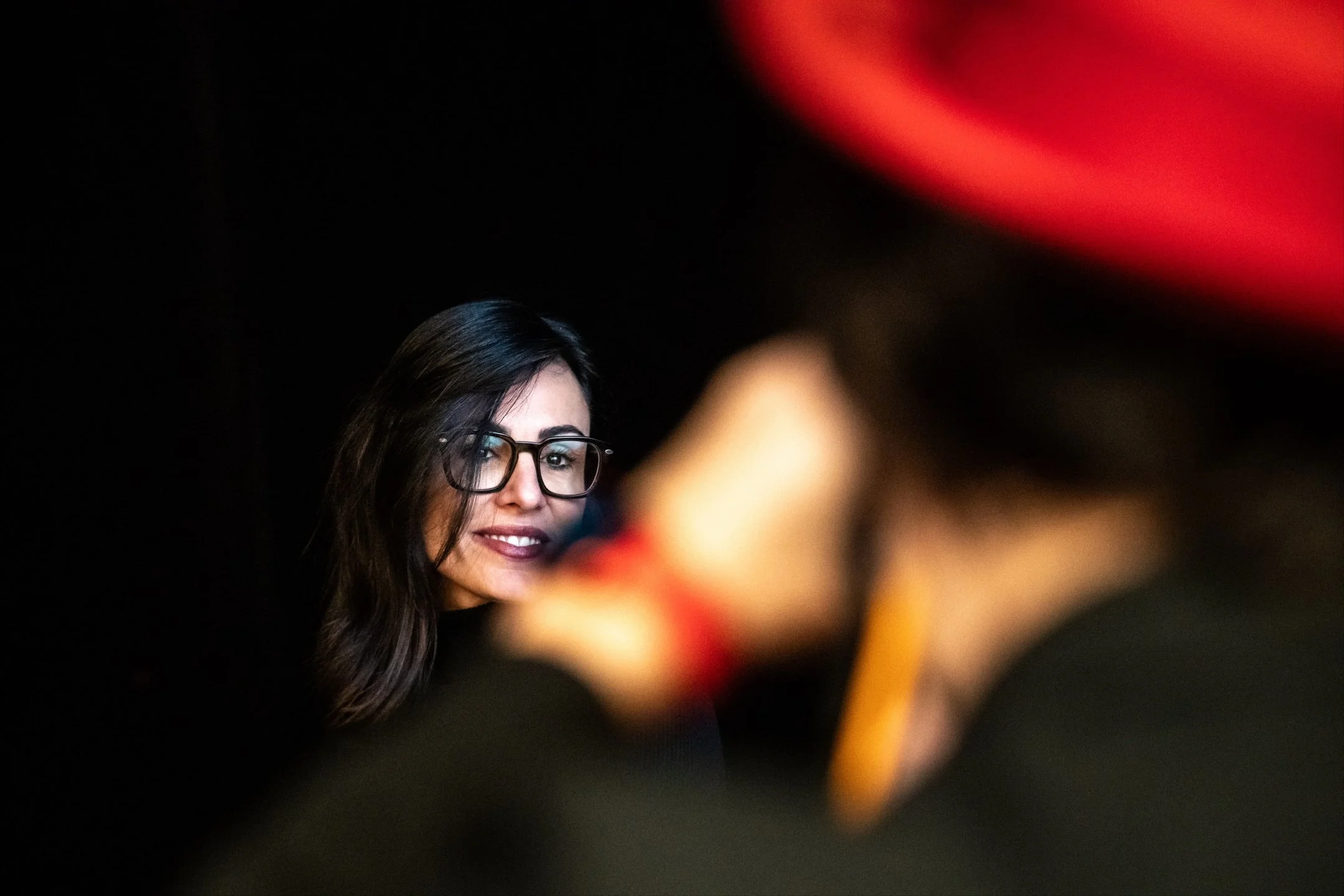 A woman with dark hair, glasses, and makeup looking or smiling at the camera, with a blurred figure in the foreground.