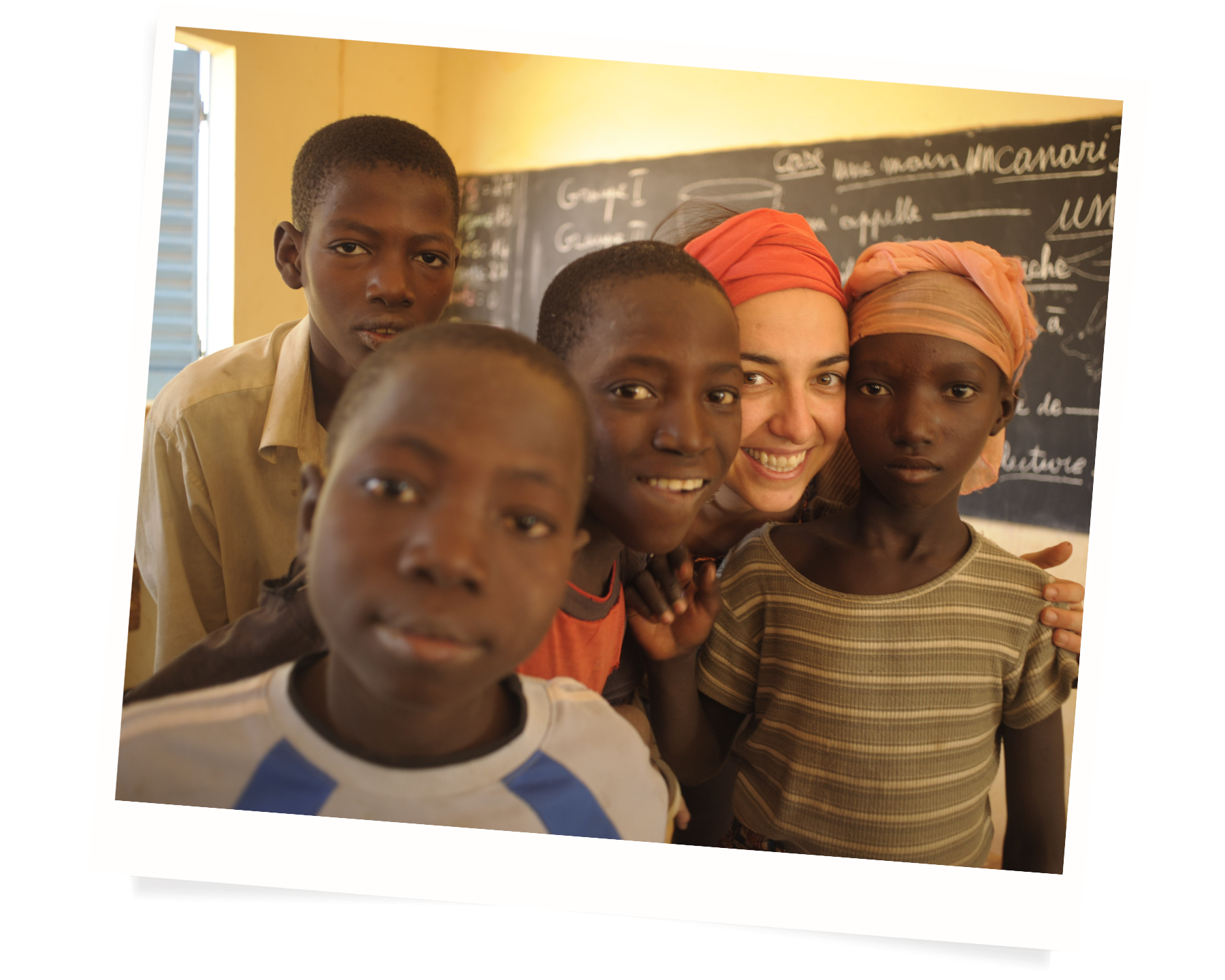 A diverse group of five children and Elena smiling together in a classroom, with a chalkboard in the background.