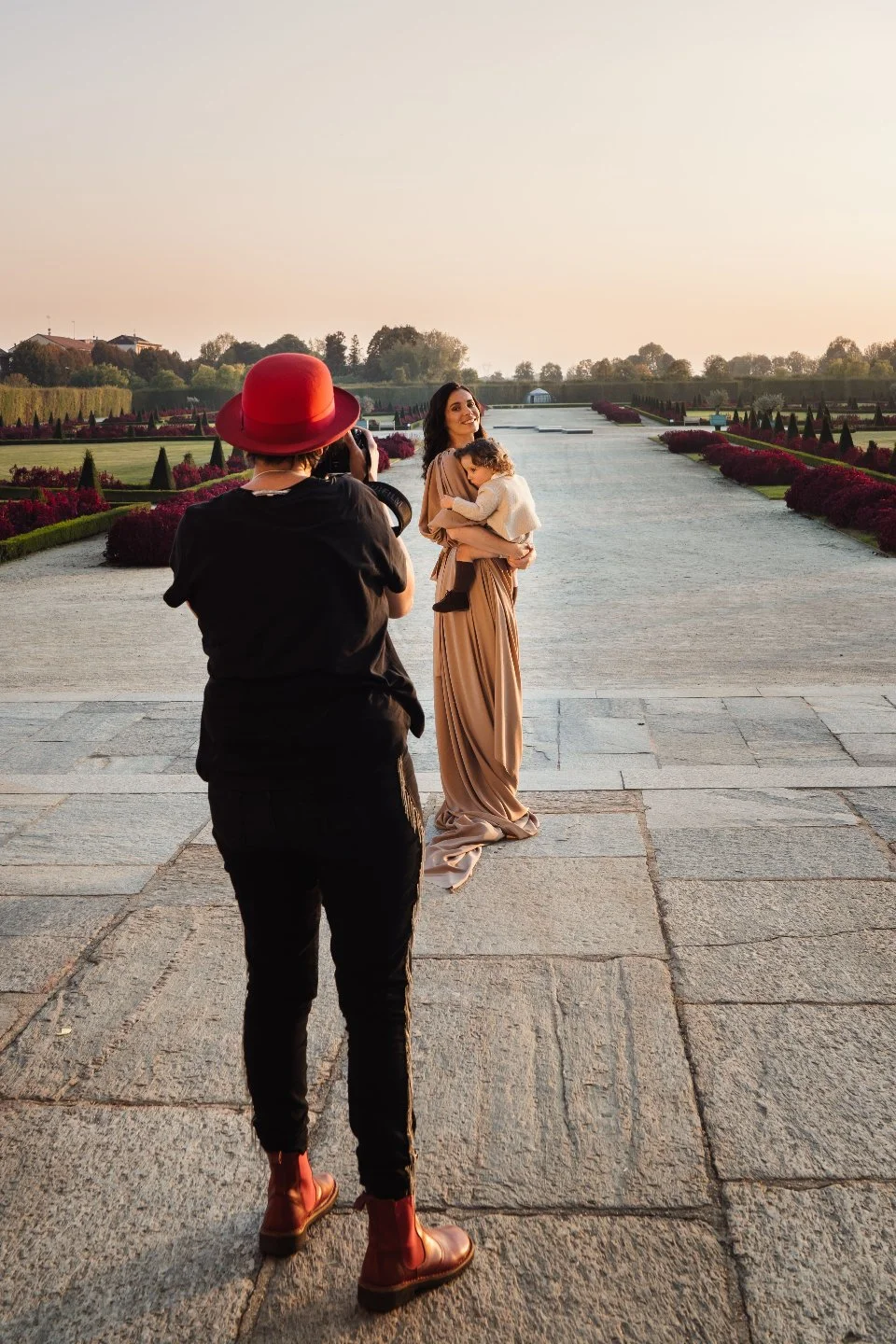 A woman holding a child standing on a pathway in a park or garden, being photographed by a person wearing a red hat and black clothing during sunset.