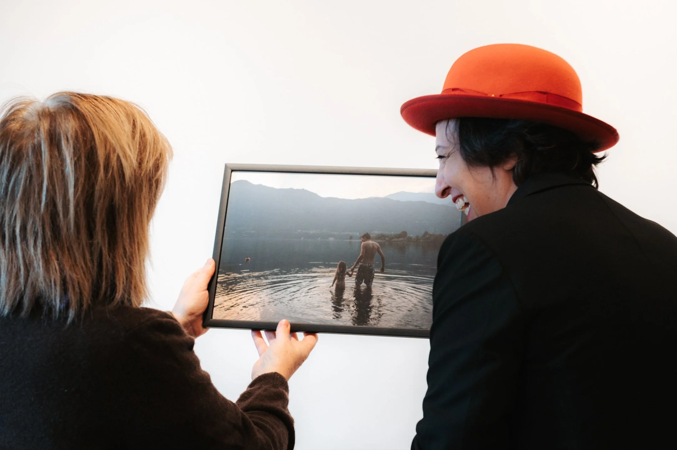 Two women are looking at a framed photograph of a man and girl swimming in a lake with mountains in the background. One woman is holding the picture, and both are smiling.