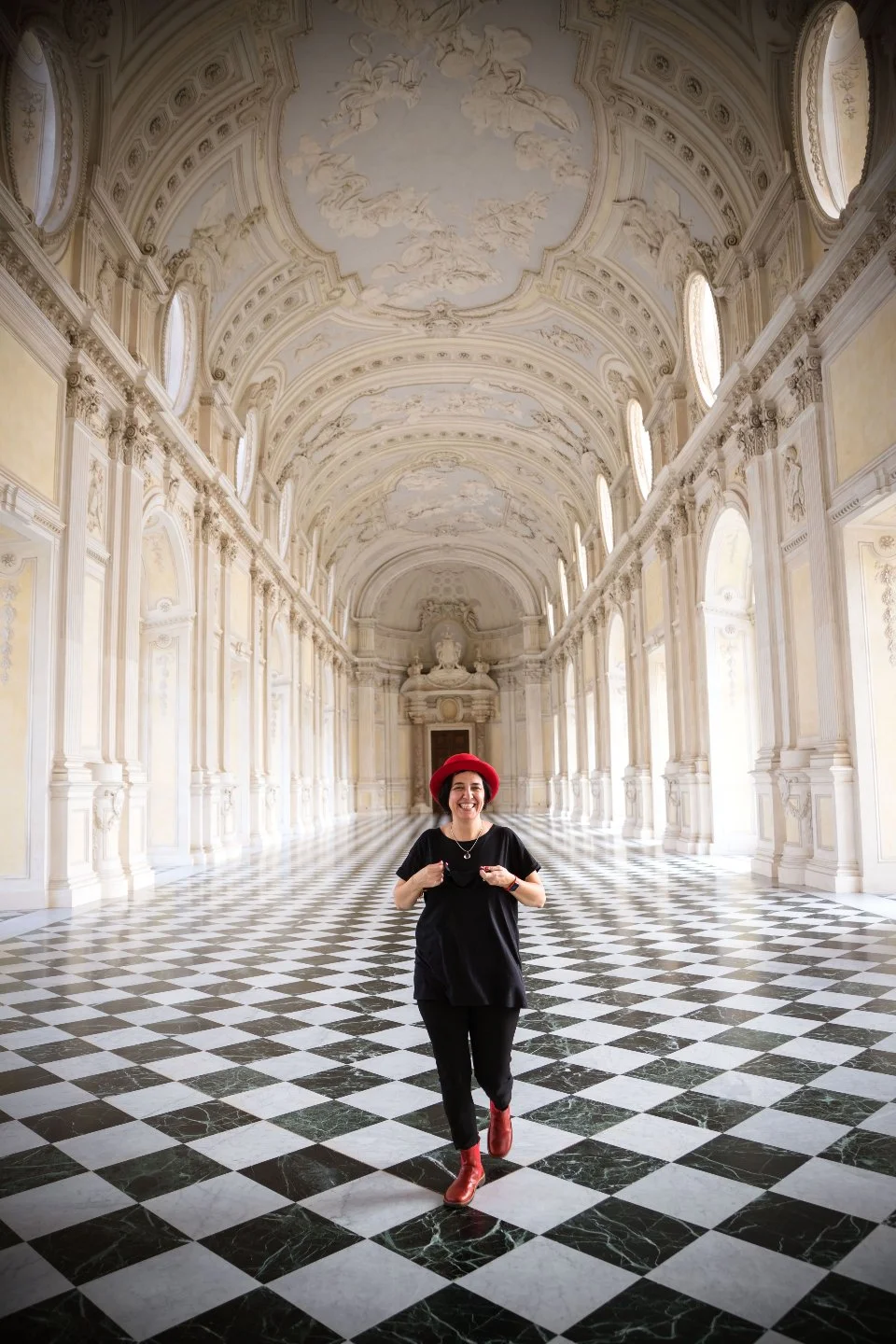 A woman in a black outfit, red hat, and red boots standing in a grand, ornate hall with high, decorated ceilings, tall windows, and a black-and-white checkered marble floor.