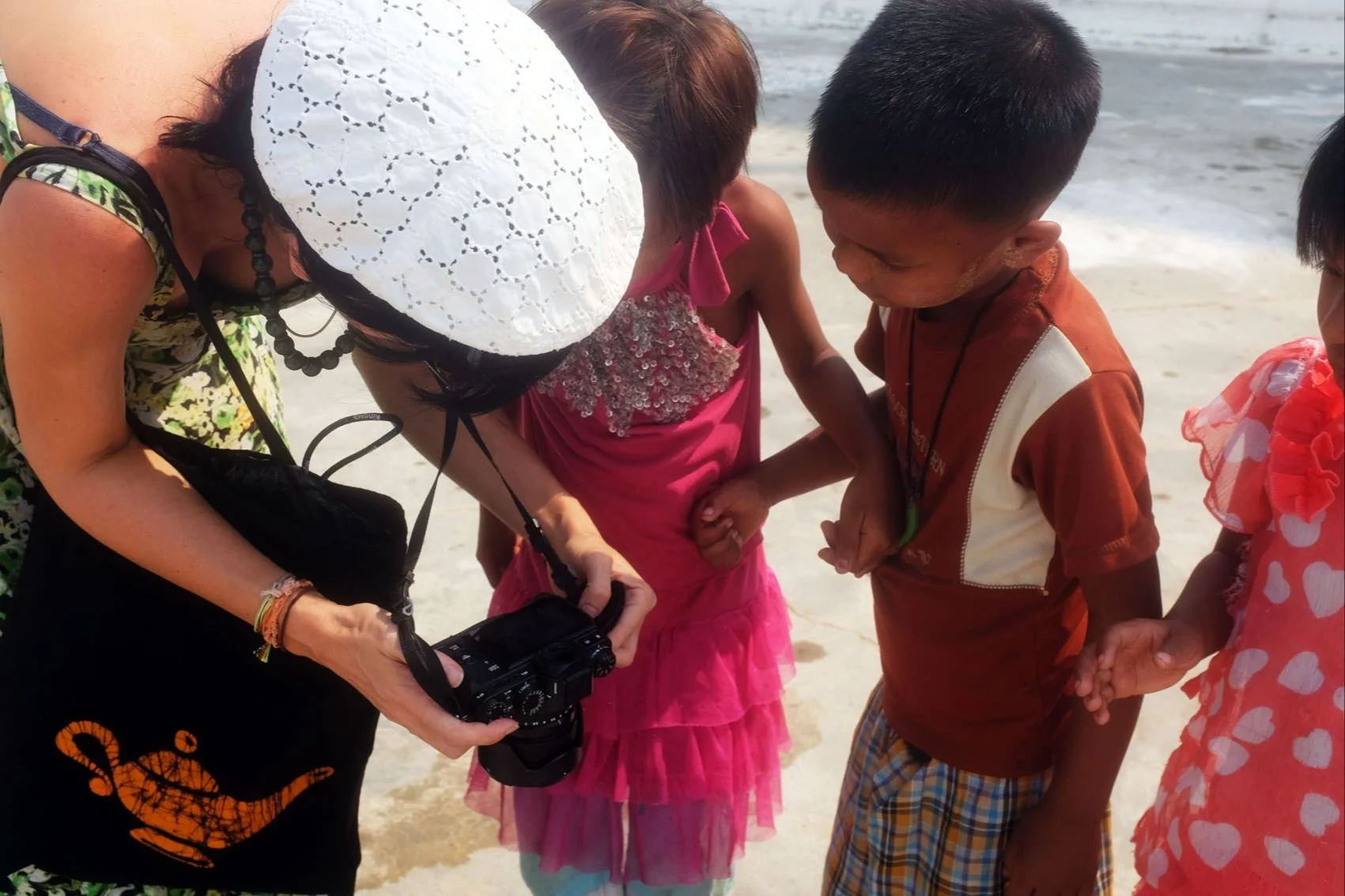 Woman with a camera, wearing a white hat, photographing children on the beach.