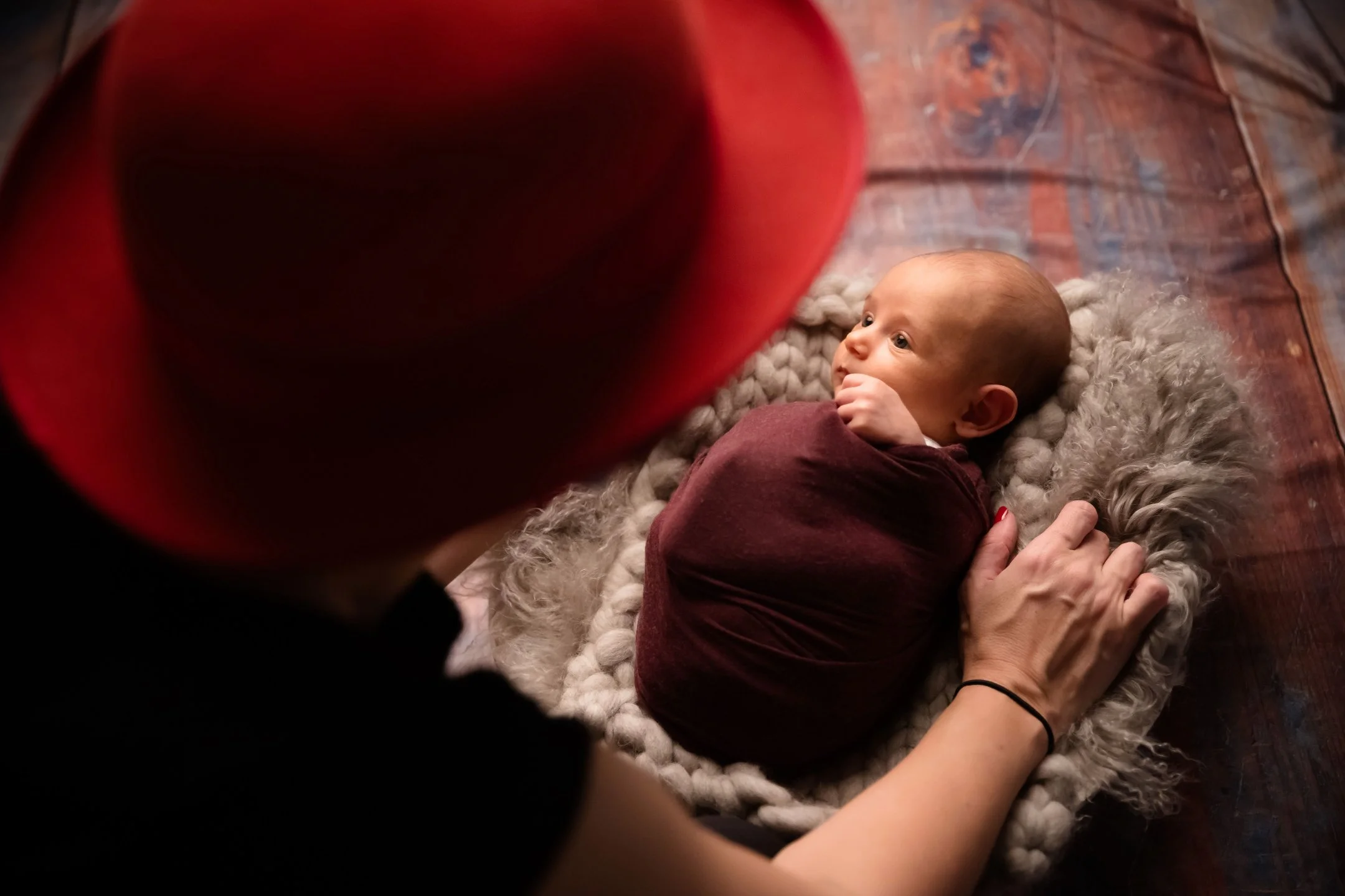 A person with a red hat and black shirt looking at a baby lying on a fluffy mat on a wooden floor. The baby is wrapped in a maroon blanket and gazes upwards with a curious expression.