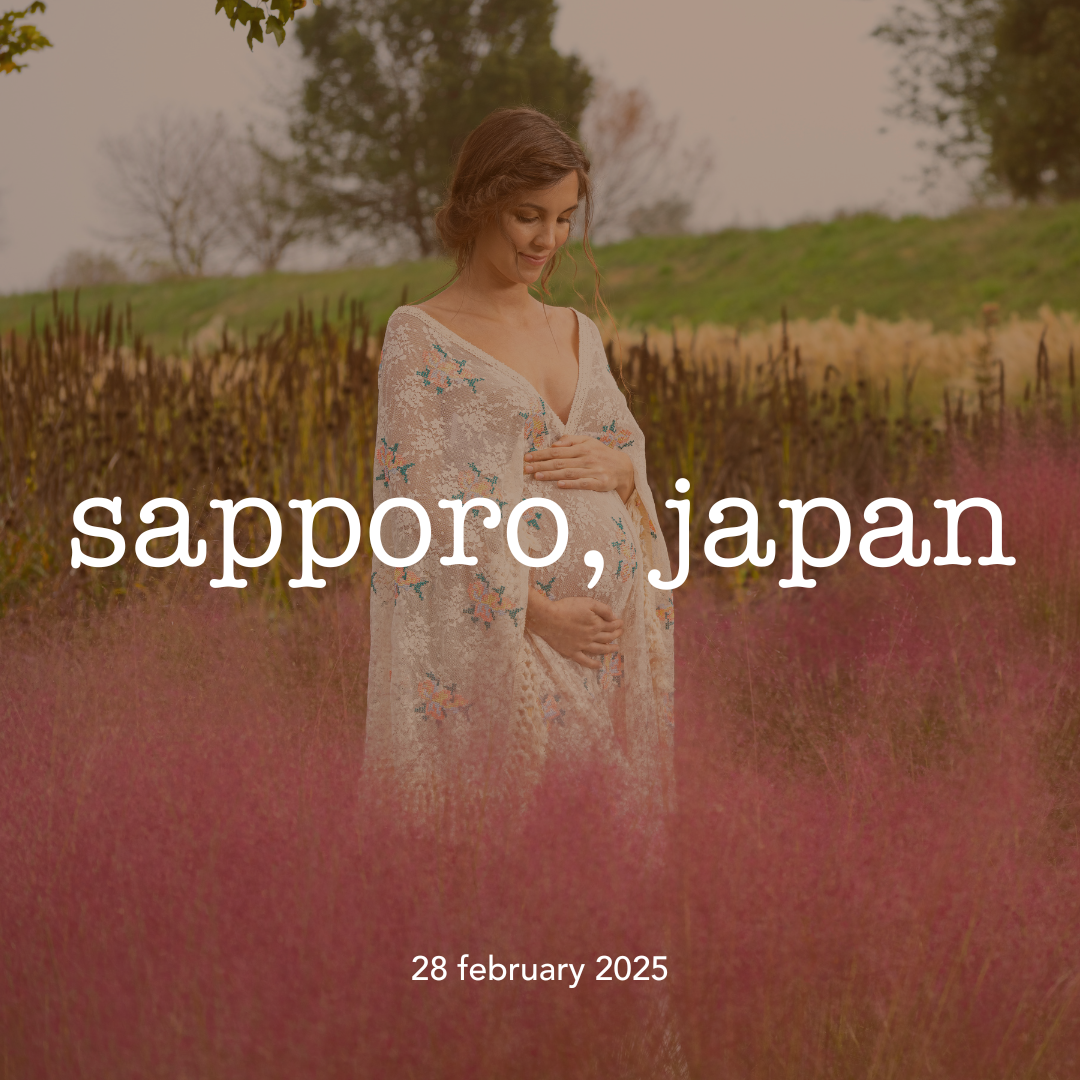 A woman standing in a field of pink flowers in Sapporo, Japan, during daytime. She is wearing a floral kimono and looking down with her hand on her belly, smiling softly.