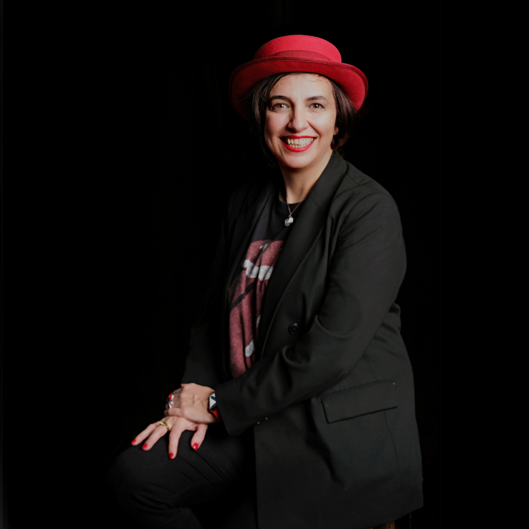 Elena, a woman with short dark hair wearing a red hat, black jacket, and graphic T-shirt, sitting against a black background, smiling at the camera.