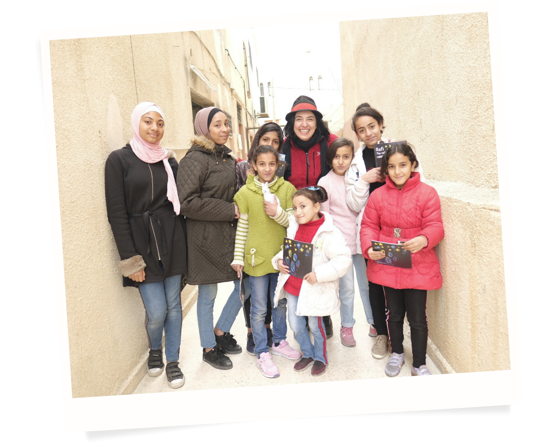 Group of children and a woman smiling in an outdoor alleyway, holding books or artwork, dressed in jackets and casual clothing.