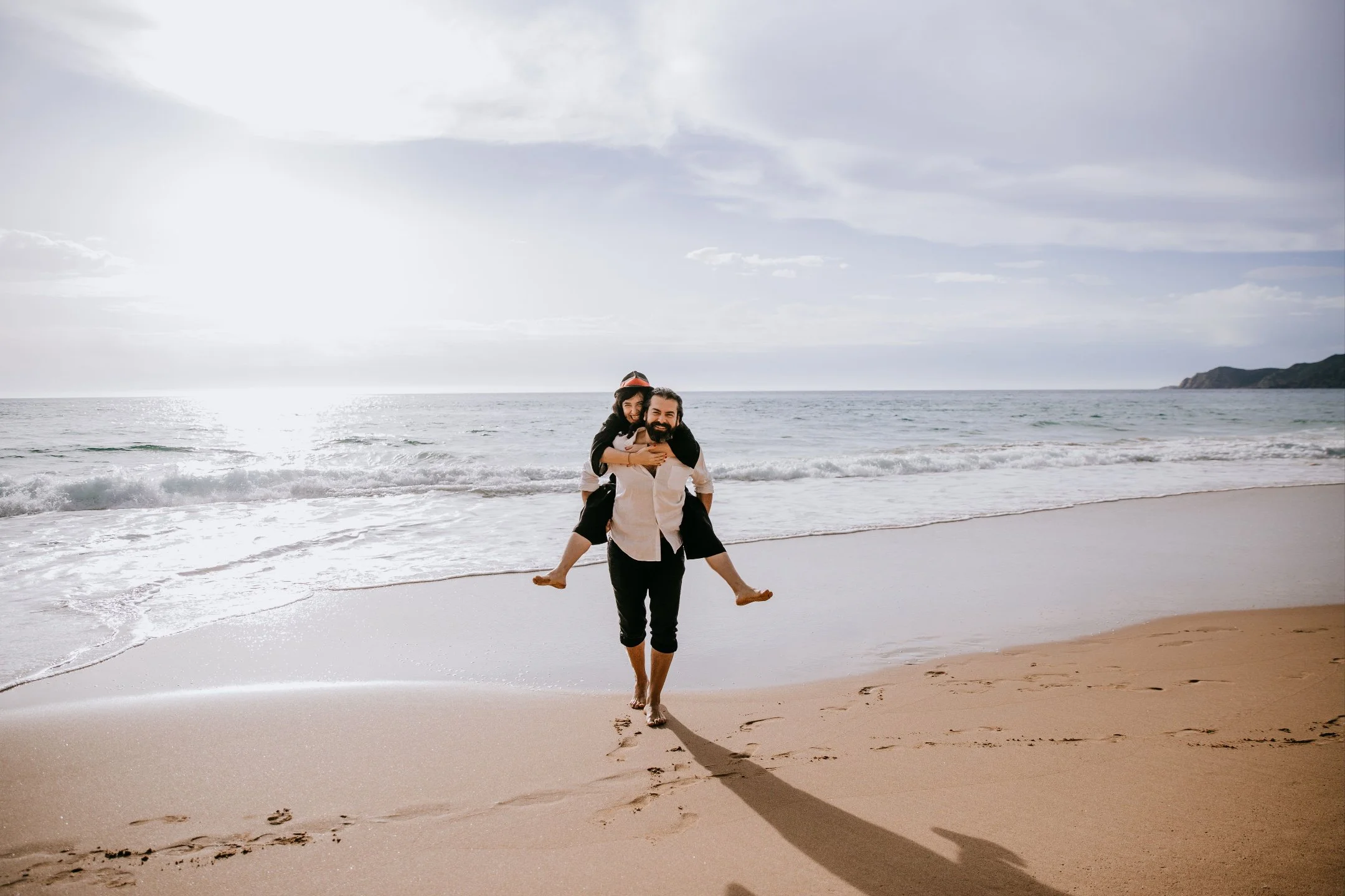 A couple walking on the beach with a woman piggybacking on a man, smiling happily near the shore with the ocean and cloudy sky in the background.