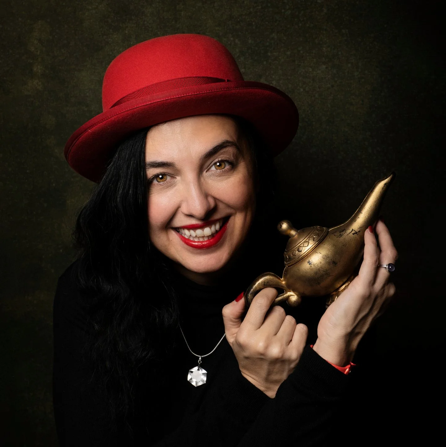 A woman with black hair, wearing a red hat, black top, and red lipstick, smiling and holding an antique brass oil lamp against a dark background.
