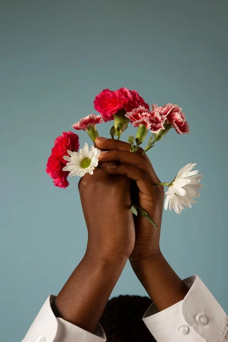 A person with dark skin holding a bouquet of pink and white carnations and daisies against a plain blue background.