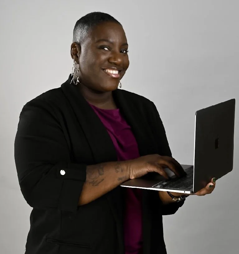 A smiling woman with short hair, earrings, and tattoos on her arm holding a laptop in front of a plain gray background.