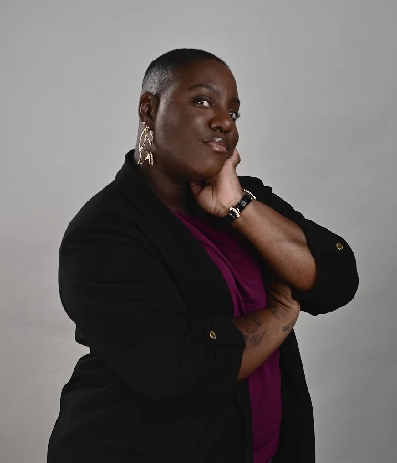 A woman with short hair, wearing earrings, a black blazer, and a purple top, posing with herHand on her chin against a plain gray background.
