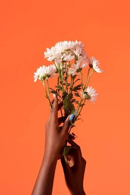 A person with dark skin holding a small bouquet of white daisies against an orange background.