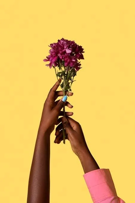 Two hands, one with light skin and one with dark skin, holding a bouquet of purple flowers against a yellow background.