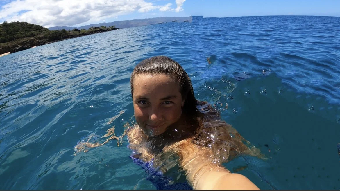 Woman swimming in the ocean, taking a selfie with a scenic coastline and mountains in the background