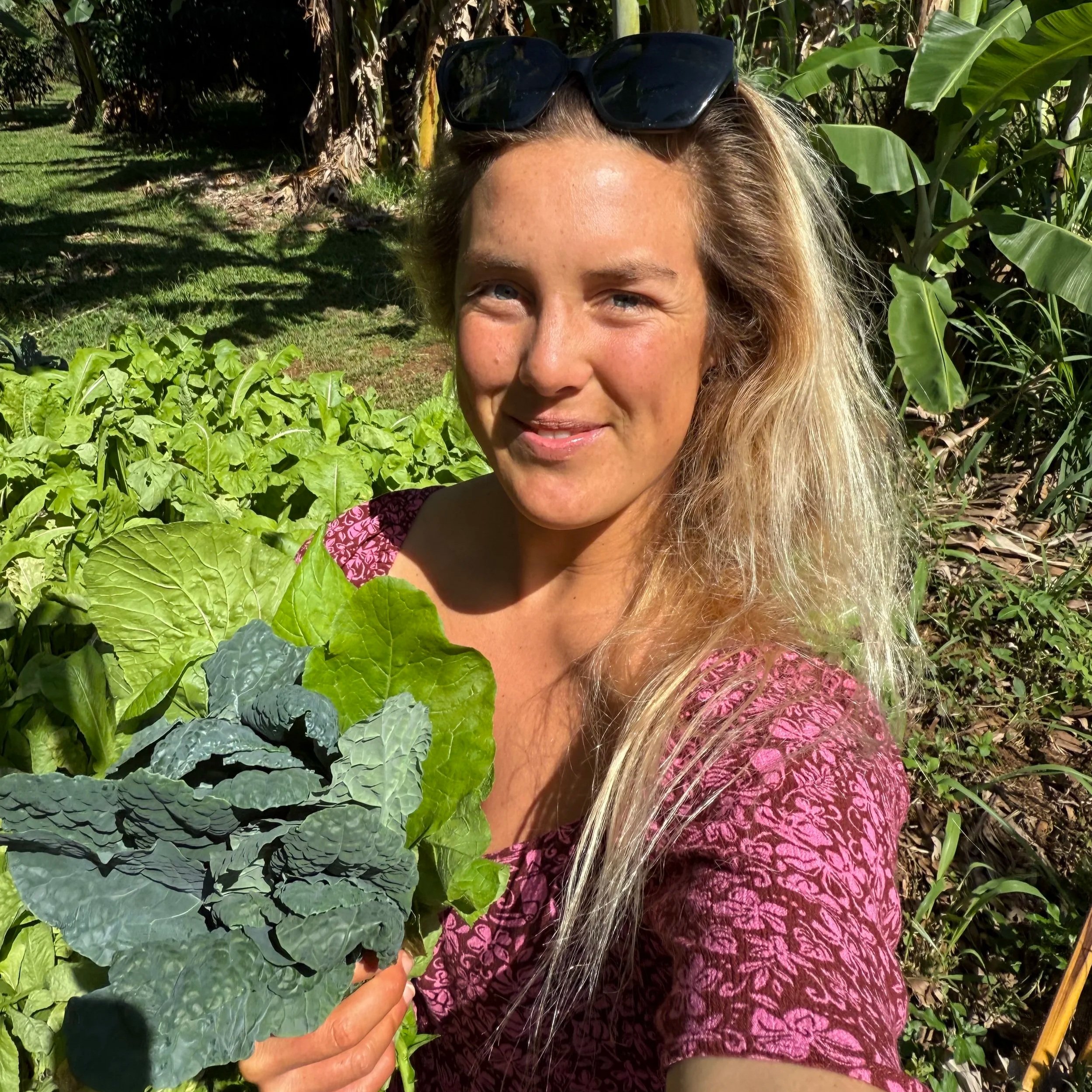 A woman with long blonde hair, wearing sunglasses on her head and a pink patterned shirt, stands outdoors in a garden holding fresh green leafy vegetables, with banana plants and other greenery in the background.