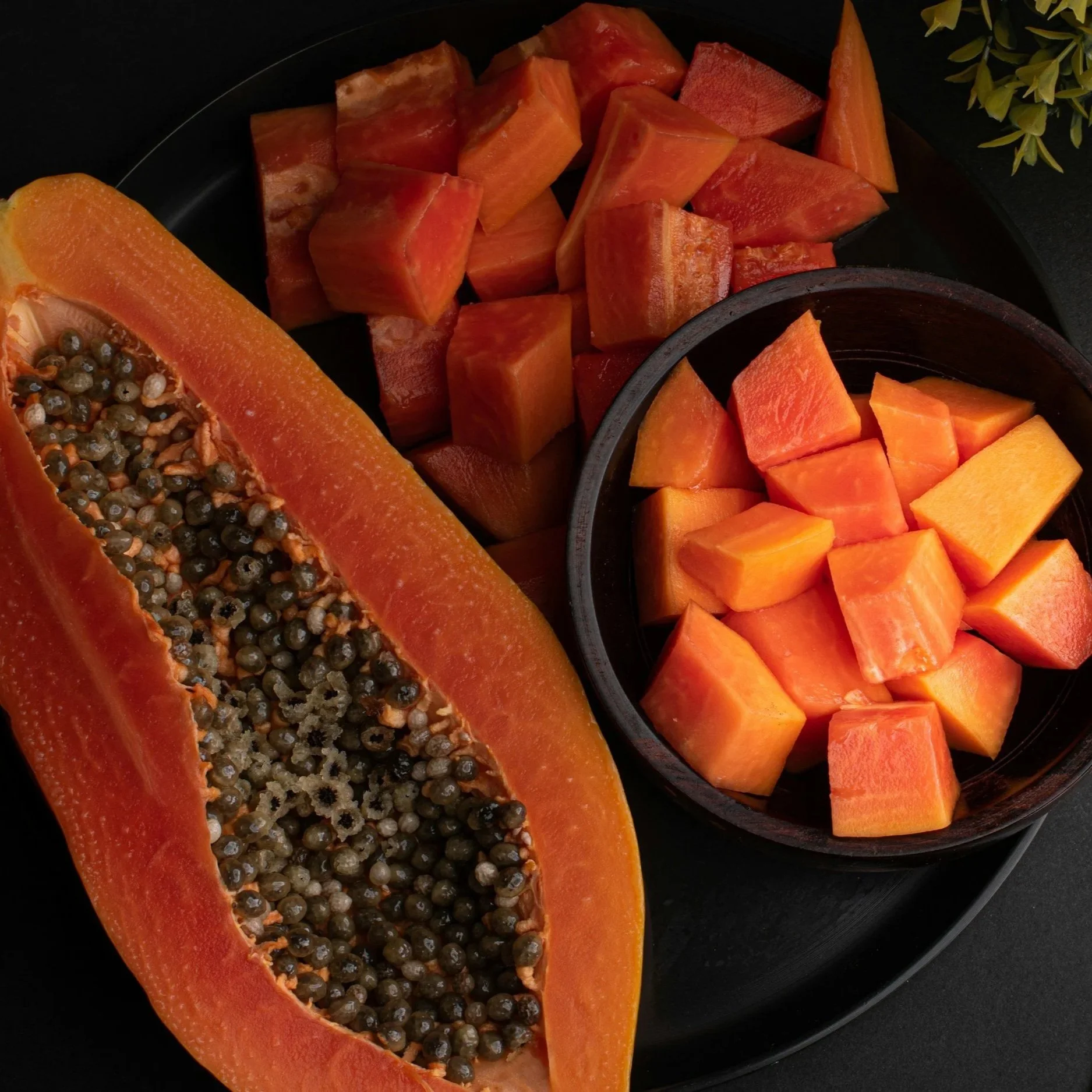 Sliced ripe papaya with black seeds, diced papaya chunks in a black bowl, and cubed papaya pieces on a black tray.
