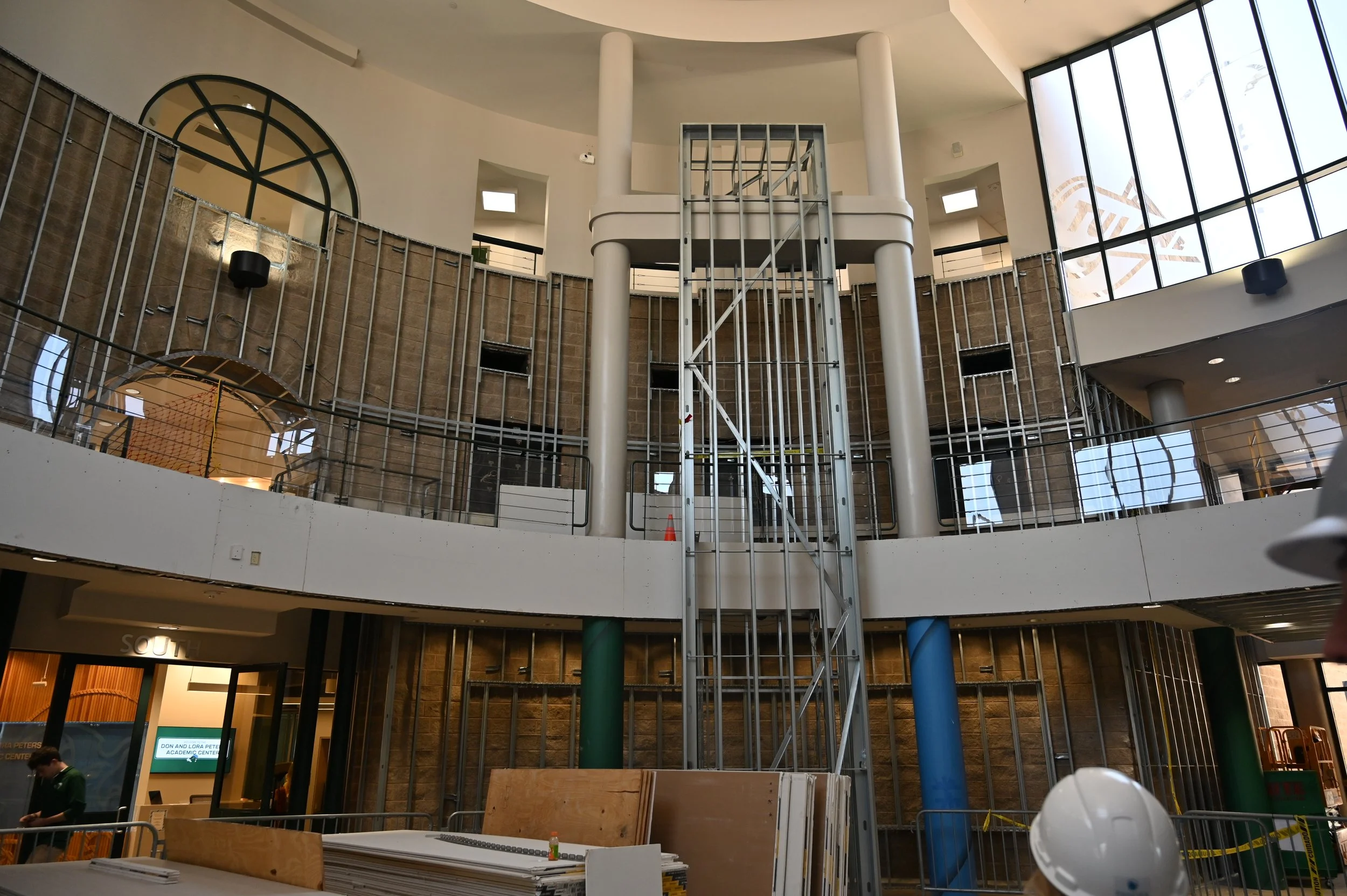 Interior of a building under construction with exposed framing, scaffolding, and large windows, and a person wearing a hard hat in the foreground.