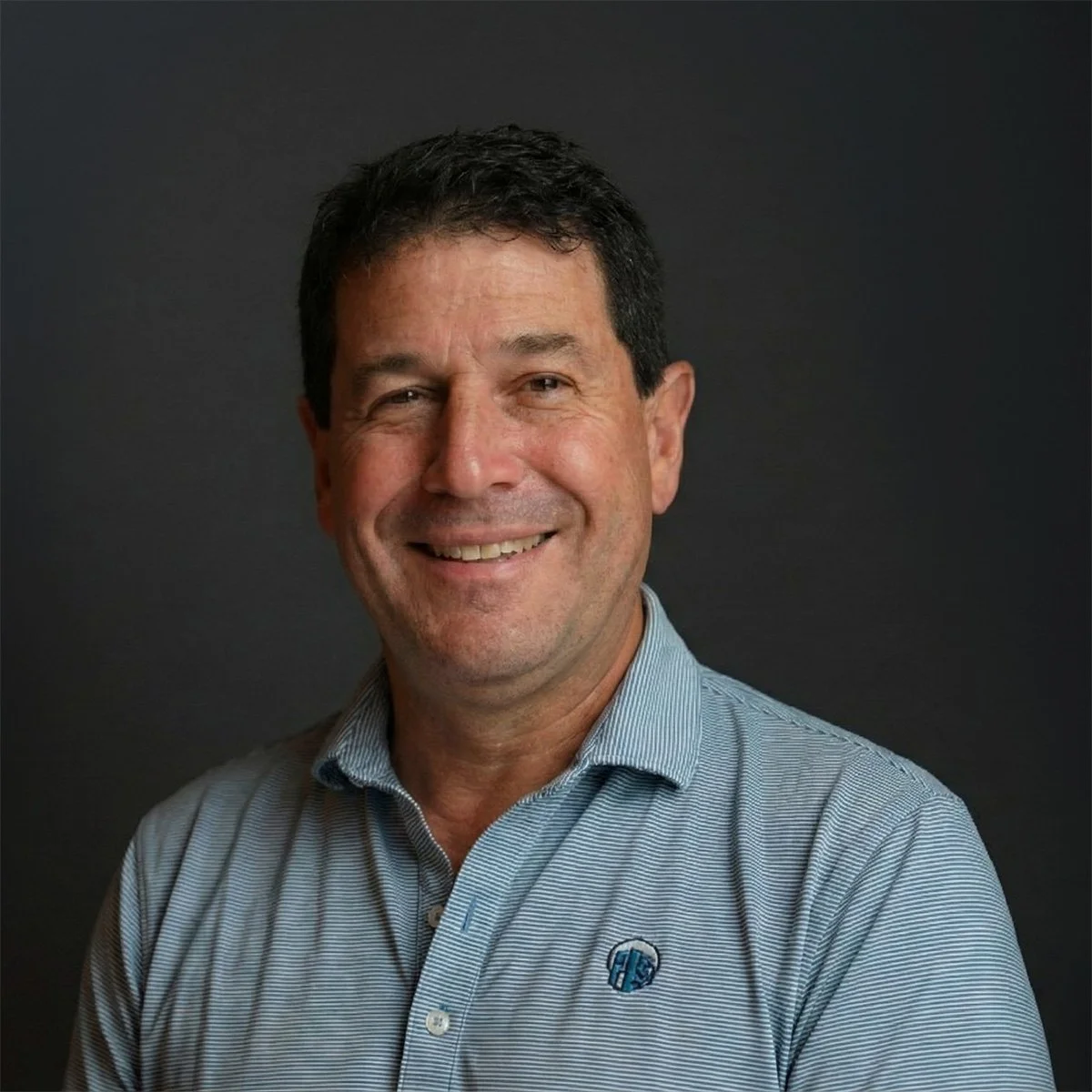 A middle-aged man with dark hair, smiling, wearing a light blue collared shirt with a logo on it, against a dark background.