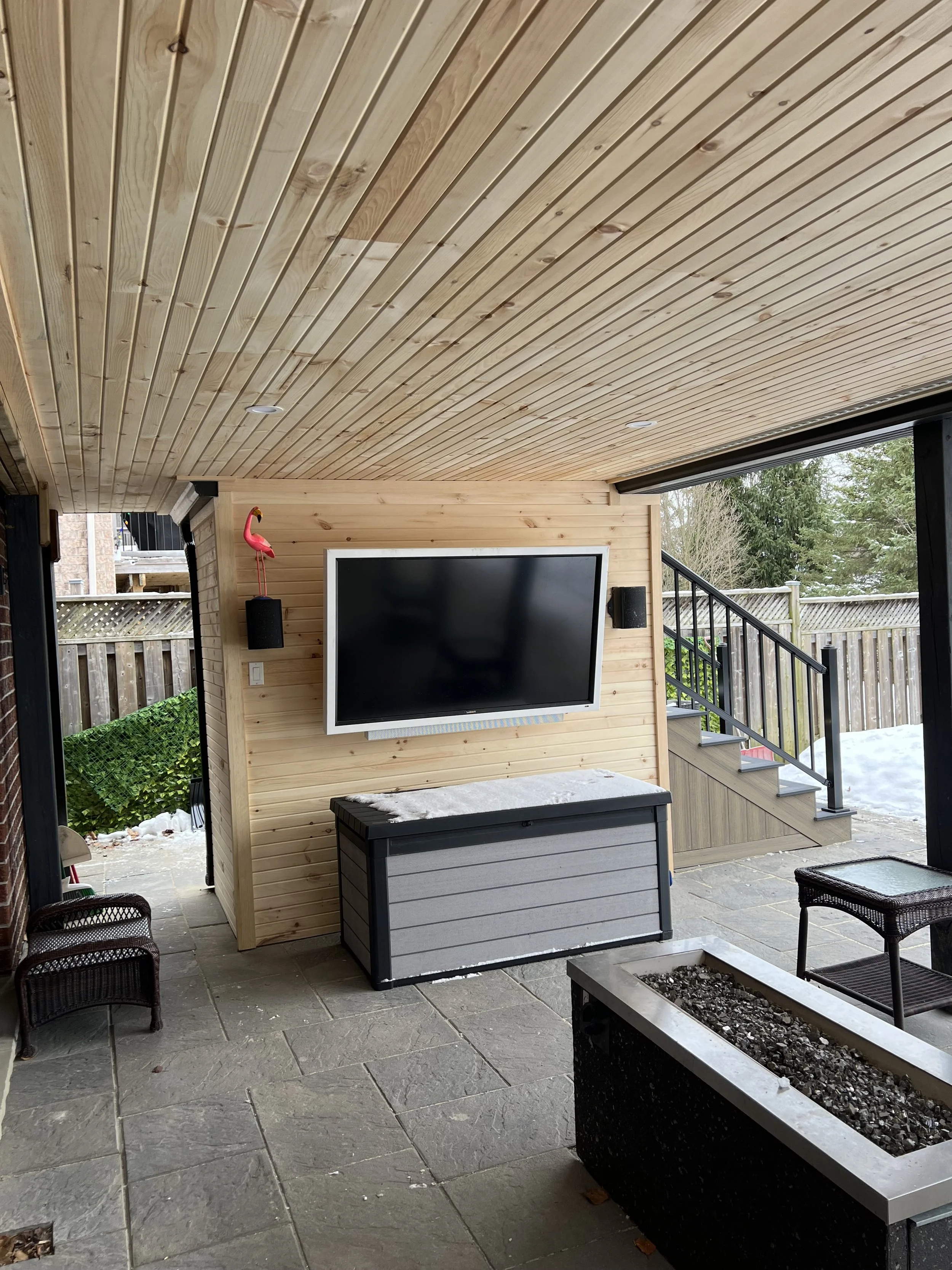 Outdoor patio area featuring a wooden ceiling, a mounted flat-screen TV, a gray storage chest, two wicker chairs, a rectangular fire pit, and a staircase leading up to a deck with snow-covered ground visible outside.