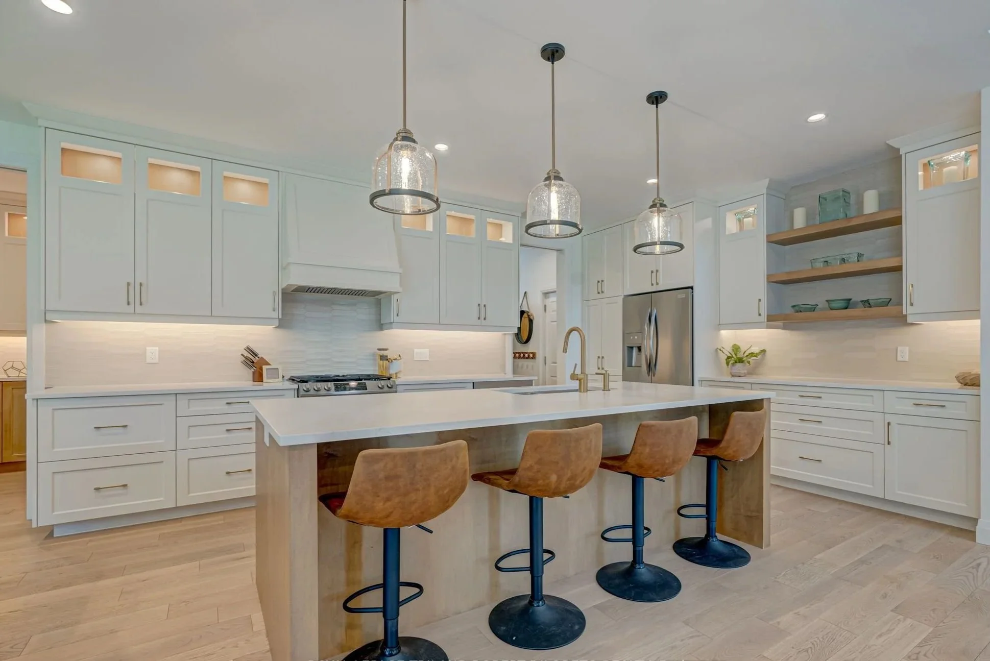 Bright, modern kitchen with white cabinetry and a large central island, featuring three brown bar stools, pendant lighting, stainless steel appliances, open shelving with dishes, and light wood flooring.