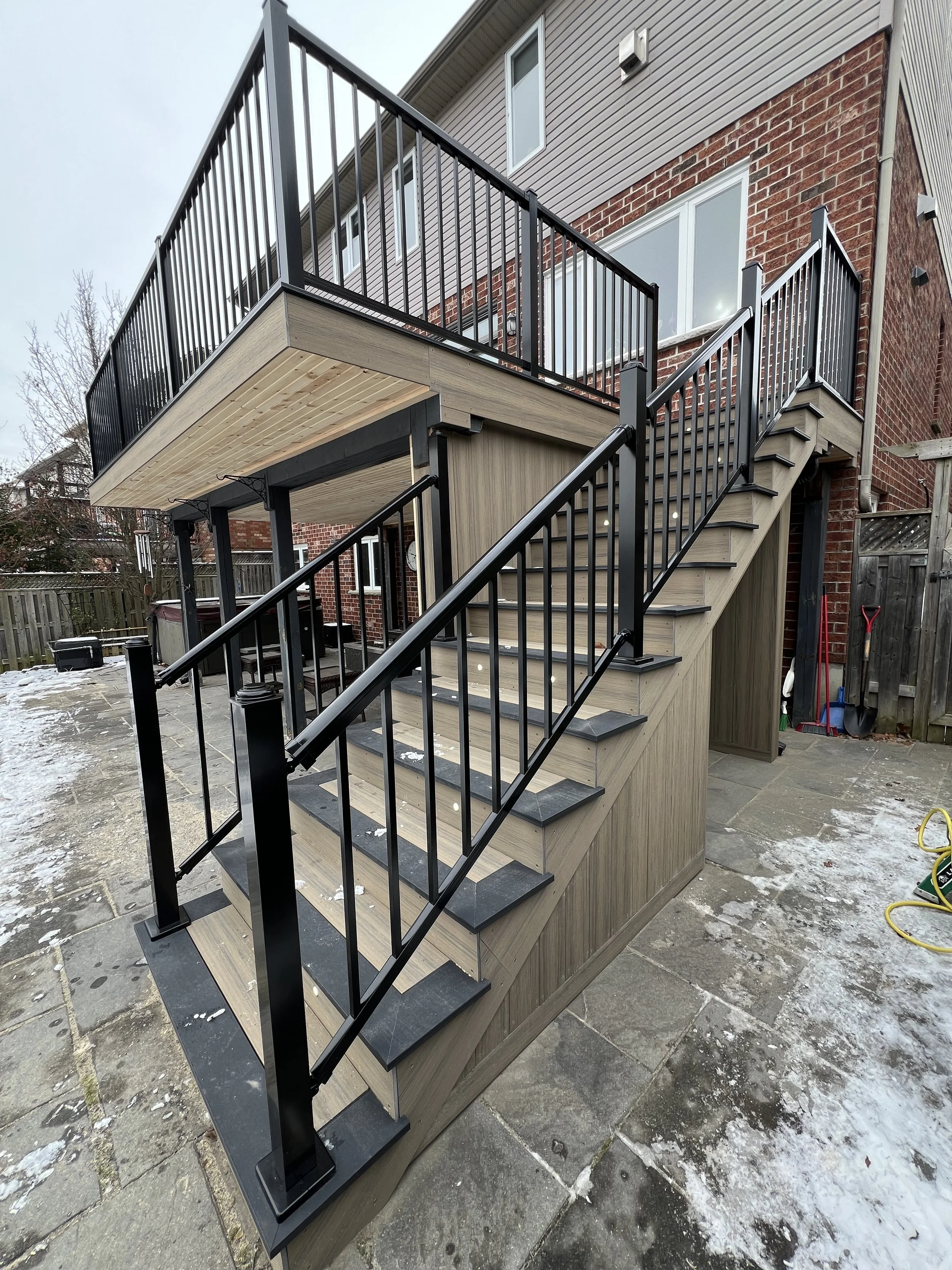 2-story composite deck renovation with staircase with black metal railings and wooden steps leading to a second-story balcony on a red brick house.