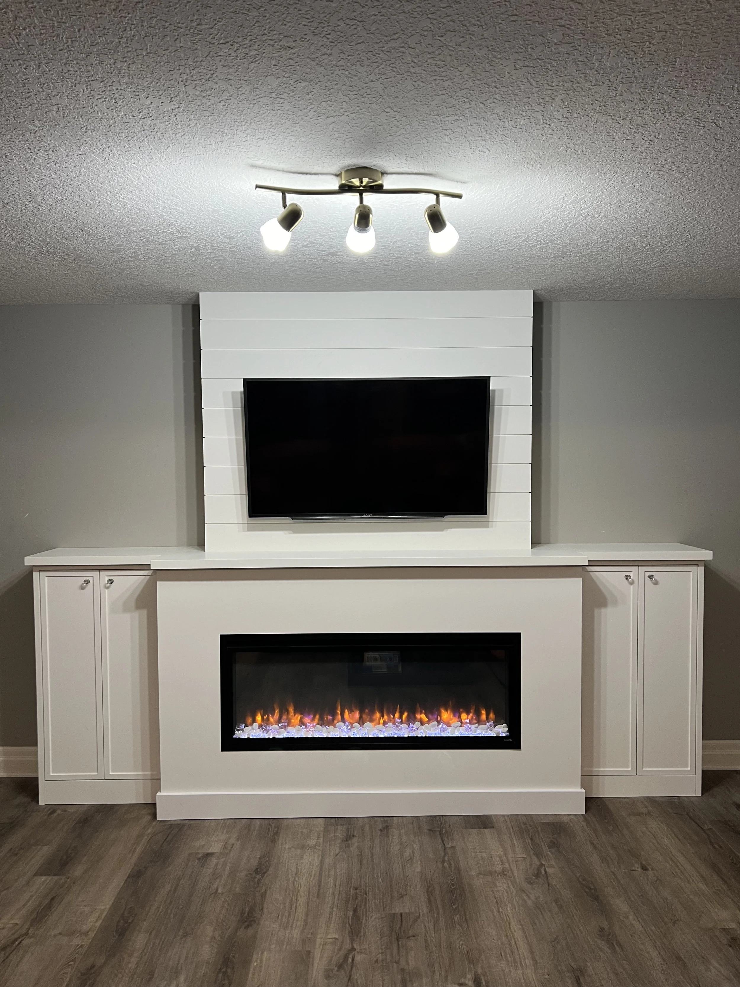 Basement renovation fireplace with a mounted flat-screen TV above it and white cabinetry on each side, with a textured ceiling and wooden floor.