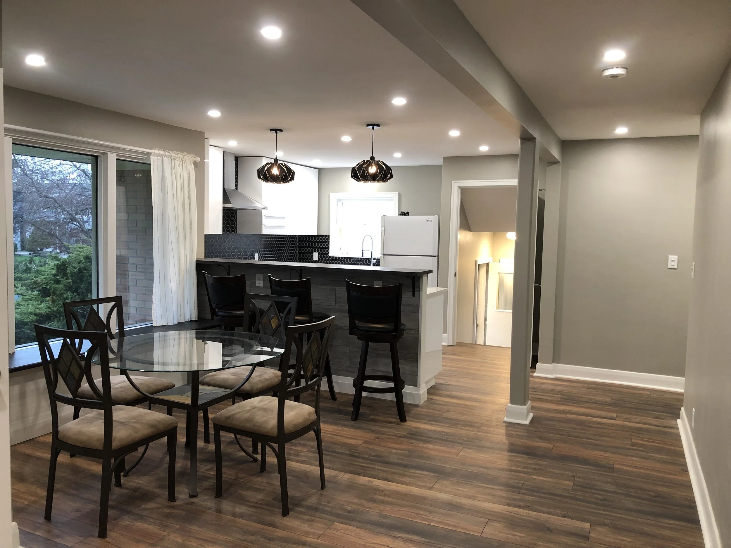 Open concept living space with dining area and kitchen in a rental property, featuring hardwood floors, a glass dining table with six chairs, and a kitchen island with three chairs, pendant lighting, black backsplash, and a large window.