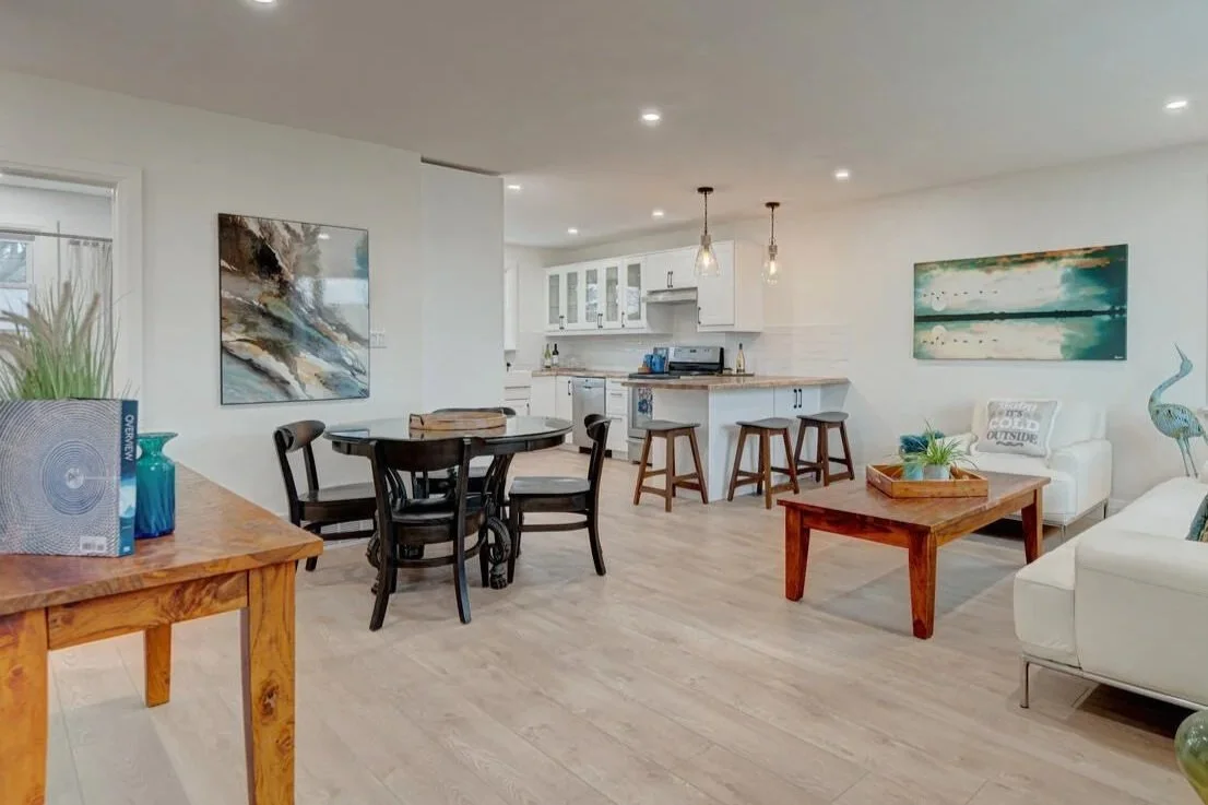 Living room with white walls, wood flooring, white kitchen with cabinets, black dining table with chairs, wooden bar stools, and modern hanging light fixtures.