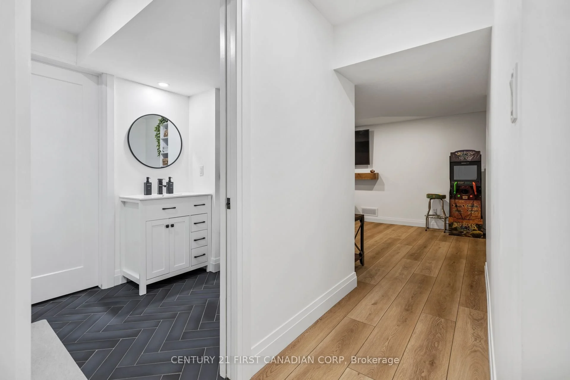 A view of a basement hallway featuring a white bathroom vanity with black hardware, black soap dispensers, a mirror, and a living area with a vintage arcade game machine, a stool, and a TV.