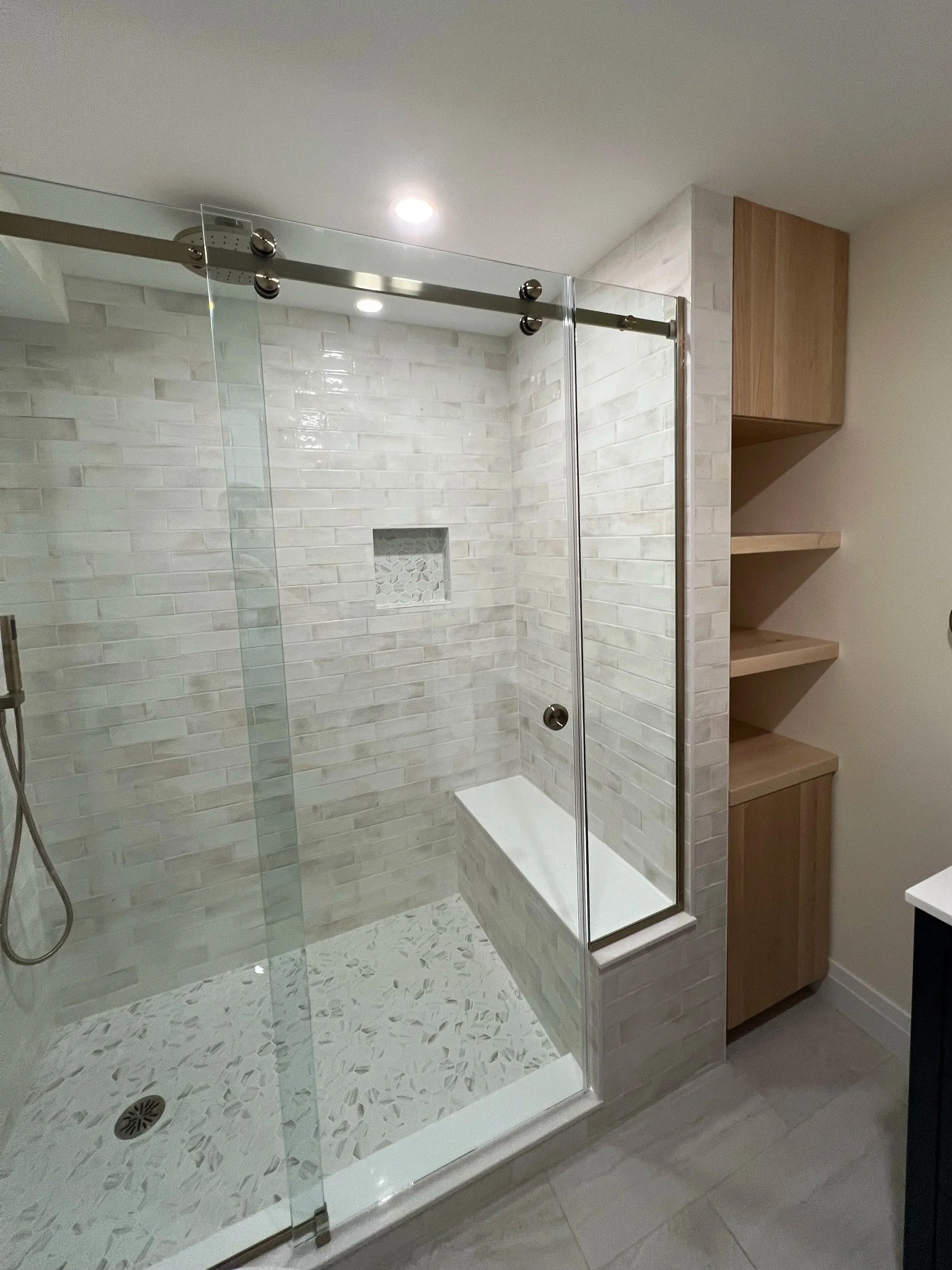 Modern bathroom shower renovation with glass doors, white tiled walls, built-in niche, a small bench, and wooden shelves nearby.