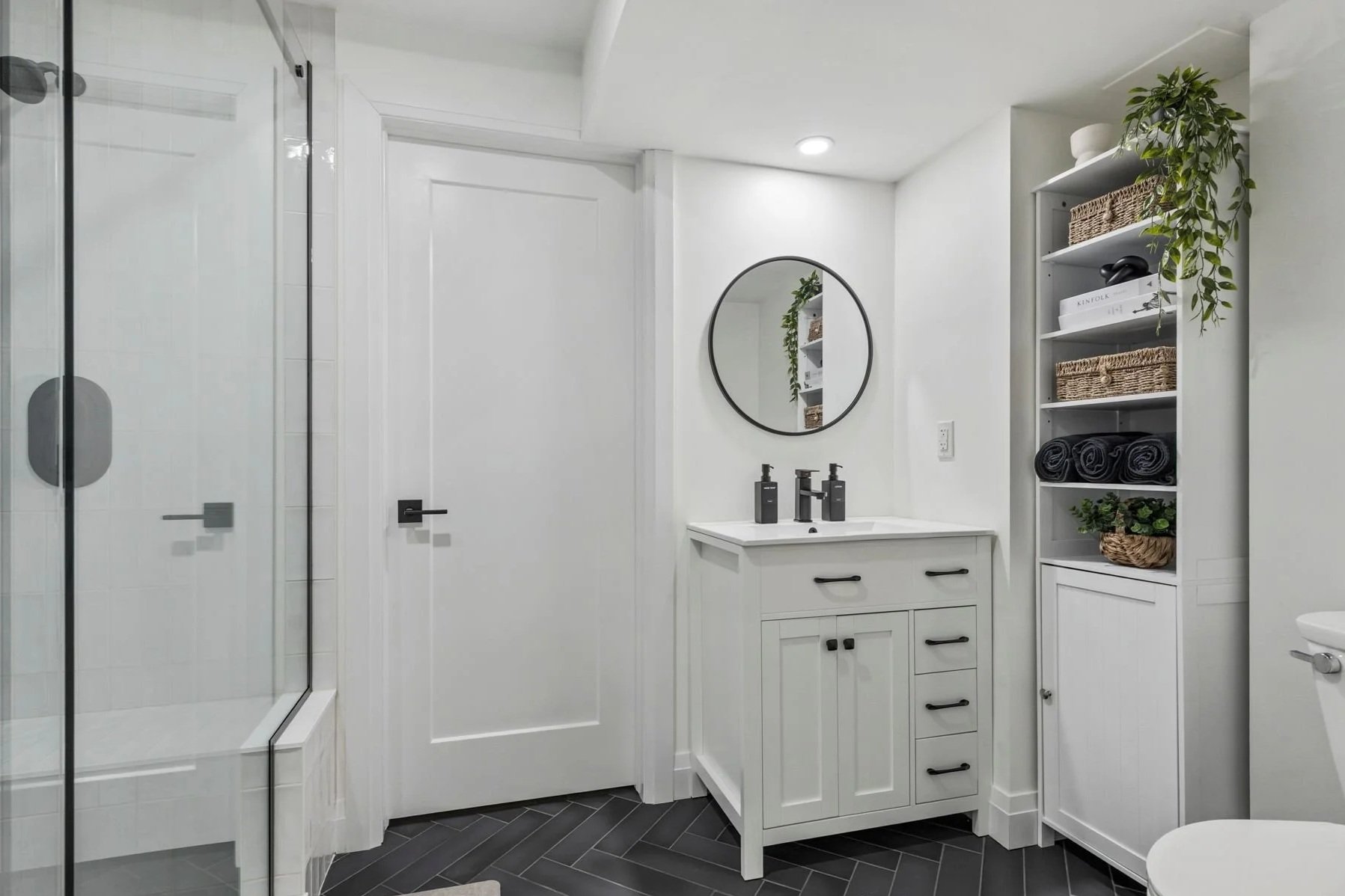 Modern white bathroom with a glass shower enclosure, white cabinetry vanity with black hardware, a round mirror, and open shelving with baskets, towels, and plants.