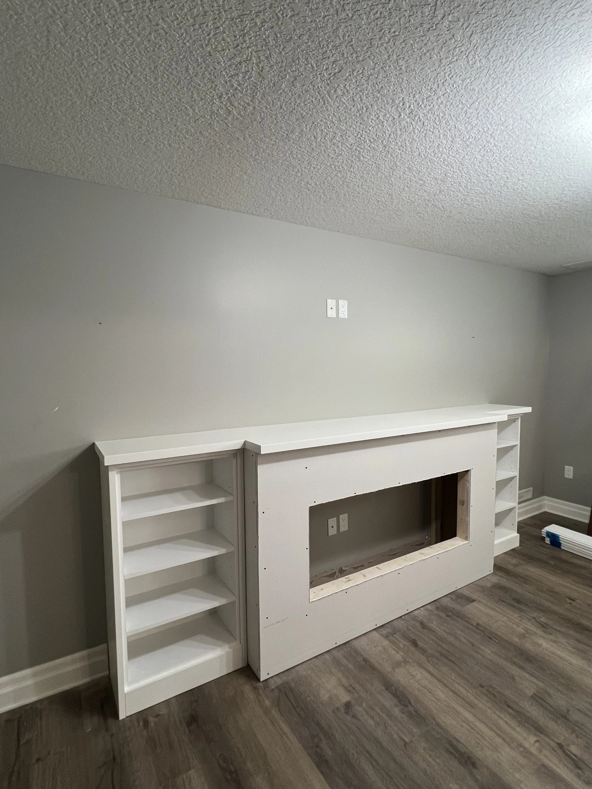 Empty living room with a white built-in fireplace mantel with open shelving on both sides, gray walls, and wood laminate flooring in a basement renovation.
