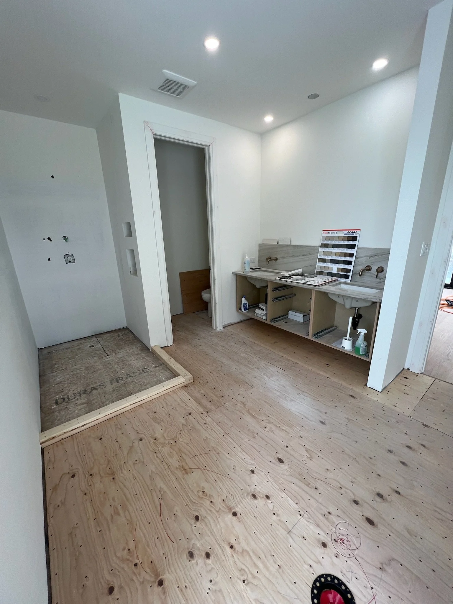 Bathroom renovation under construction with unfinished wooden flooring, a small bathroom visible through an open door, and a vanity area with a marble countertop, two sinks, and color samples on display.