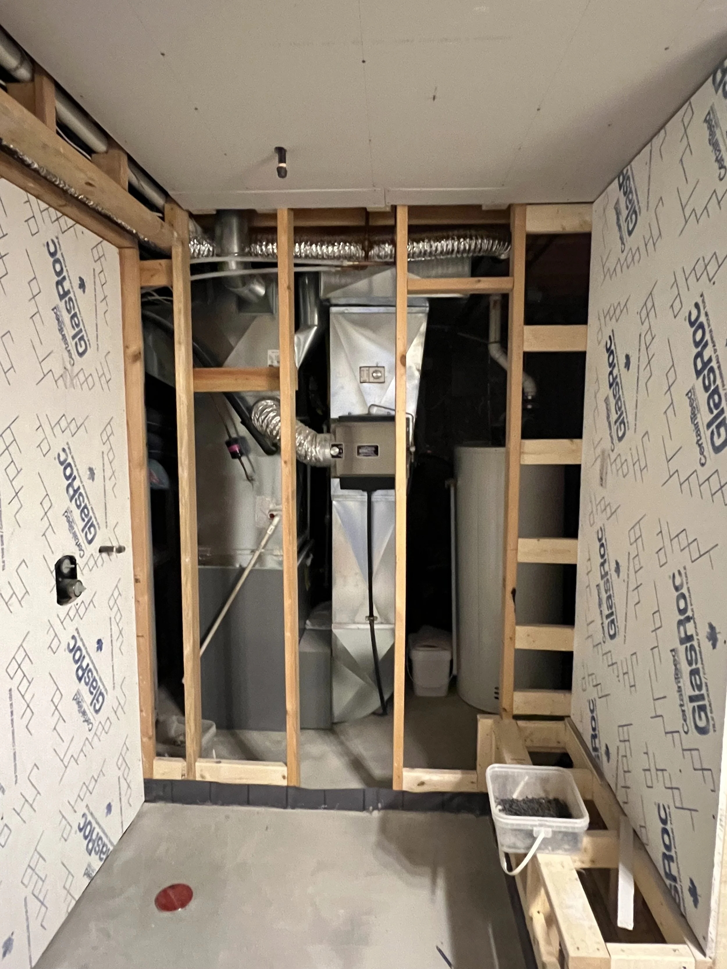View of a partially framed room with exposed wooden studs and insulation panels on the walls, showing HVAC ductwork and a water heater in the background, with construction materials on the floor.
